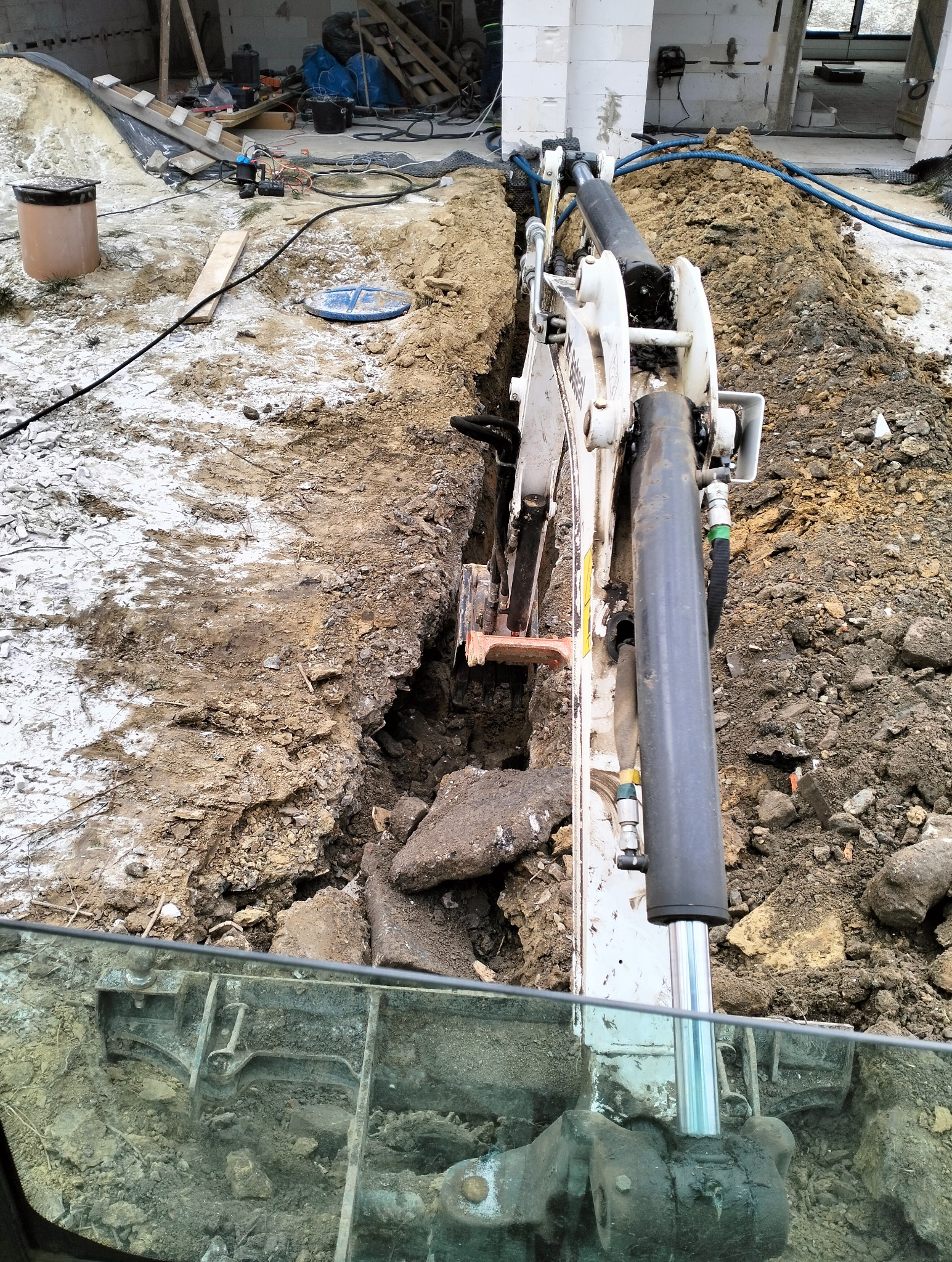 Mini koparka w trakcie wykonywania wąskiego wykopu w gruncie, widoczne fragmenty instalacji wodnej i kanalizacyjnej oraz fundamenty budynku w tle.