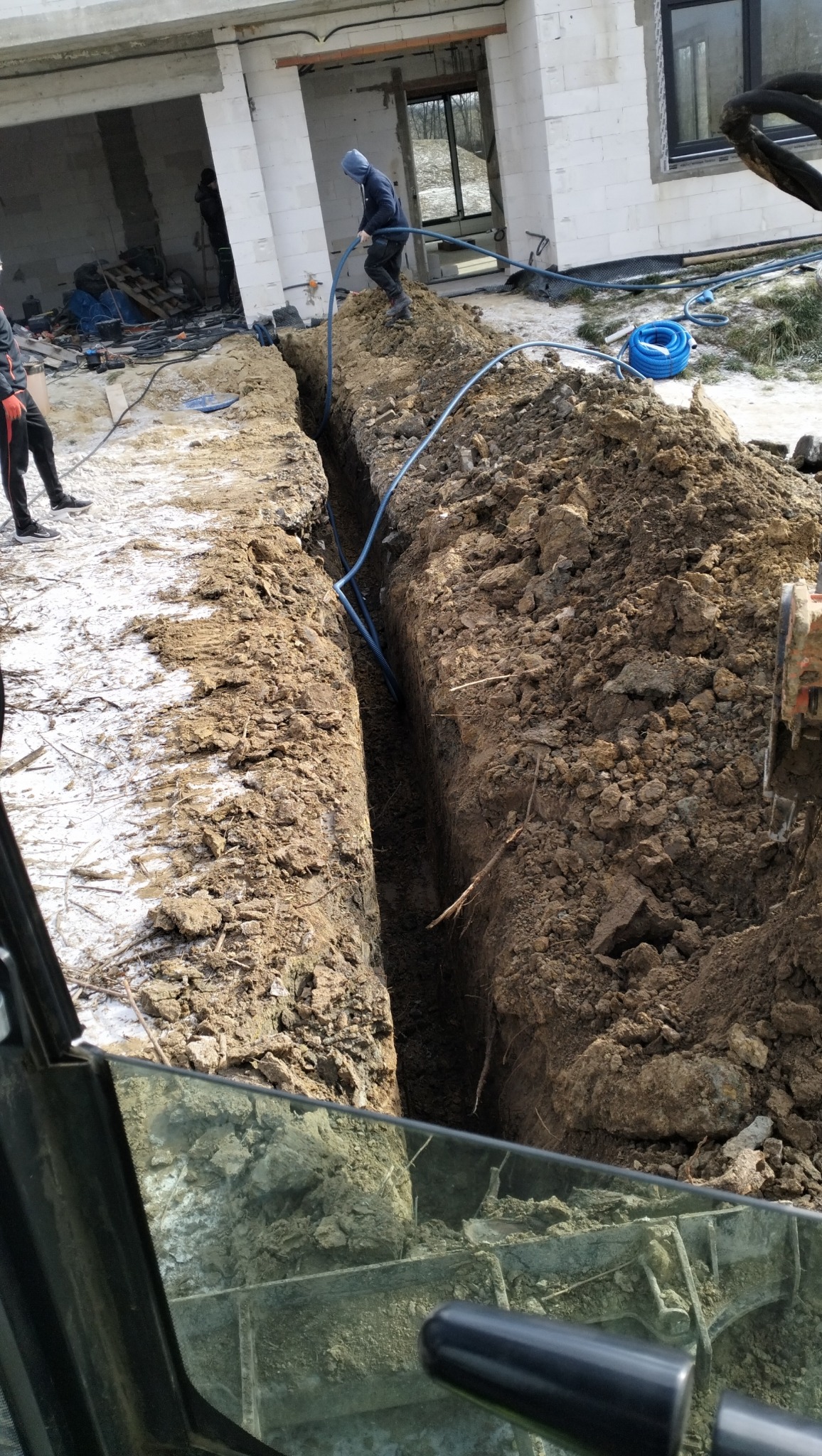 Głęboki wykop w ziemi z ułożonymi niebieskimi rurami, pracownik w bluzie z kapturem układa rury w wykopie, w tle budynek w stanie surowym z widocznymi materiałami budowlanymi.