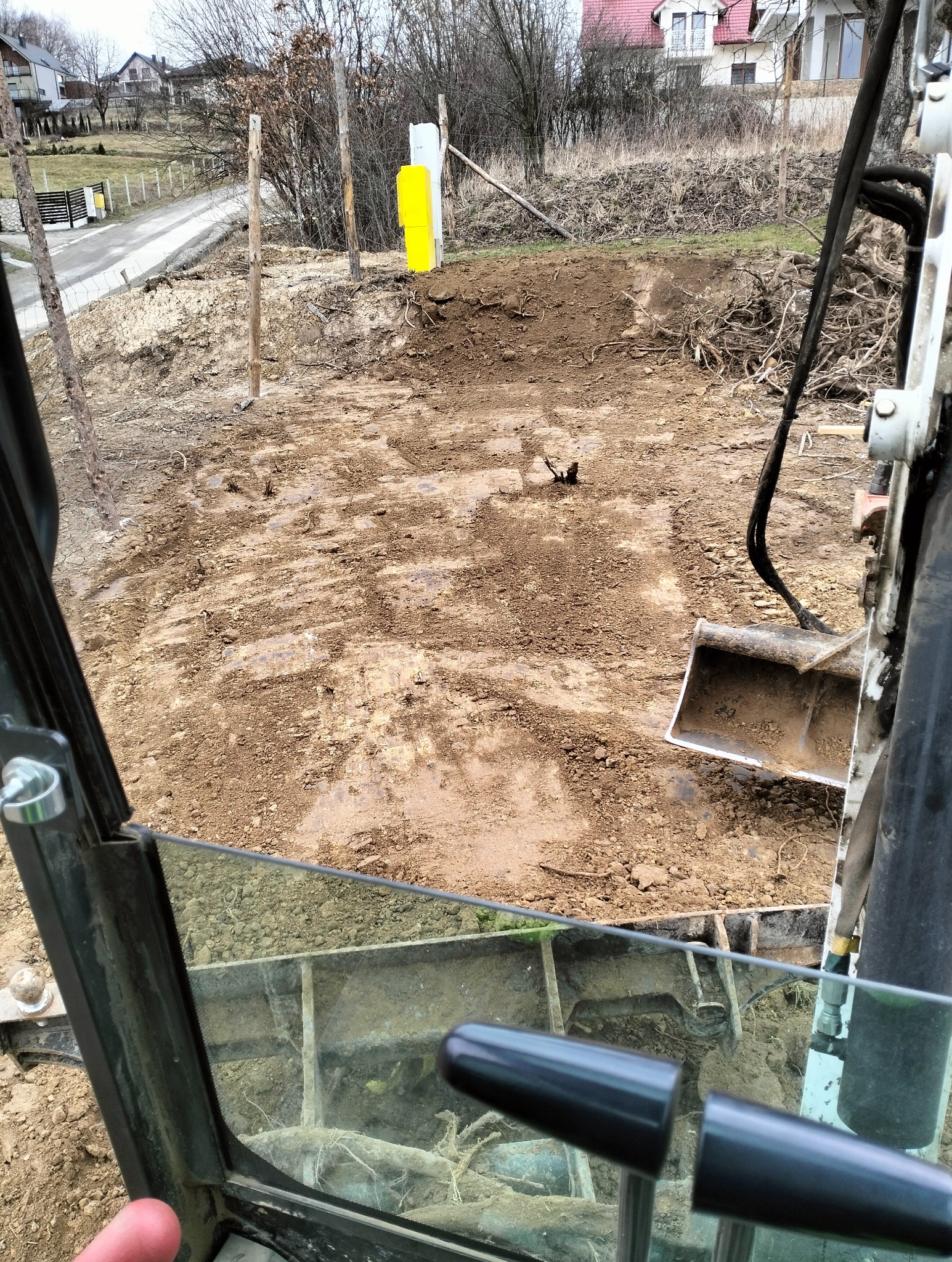Widok z kabiny koparki na teren budowy z przygotowanym wykopem, w tle widoczne drzewa i domy.