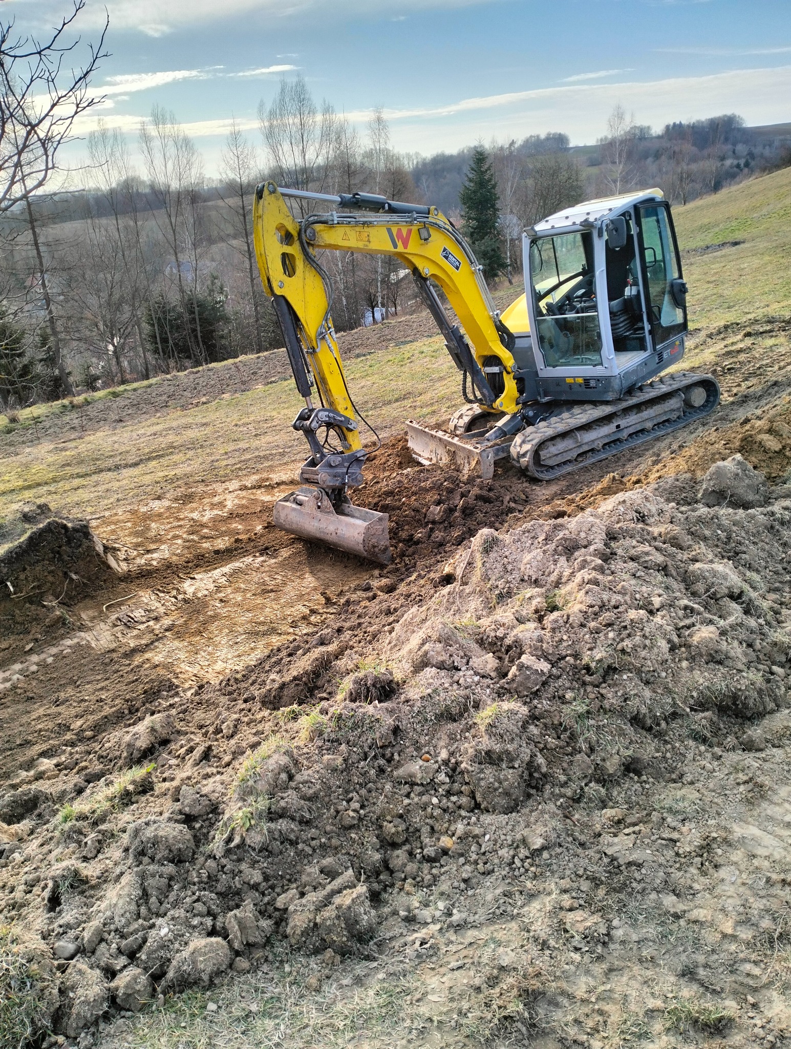 Żółta minikoparka Wacker Neuson na gąsienicach, wykonująca wykop w gruncie na pagórkowatym terenie, z widocznymi śladami pracy i stertą ziemi.