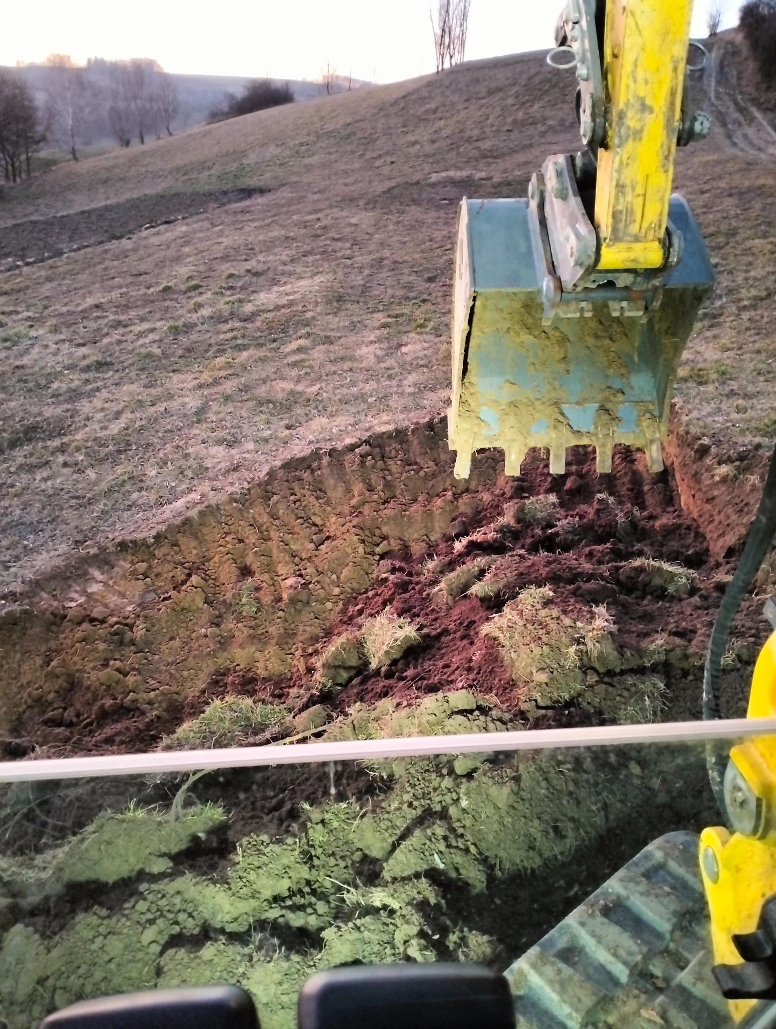 Żółta koparka z łyżką zagłębia się w wykopaną ziemię, widok z kabiny operatora na zbocze wzgórza w tle.