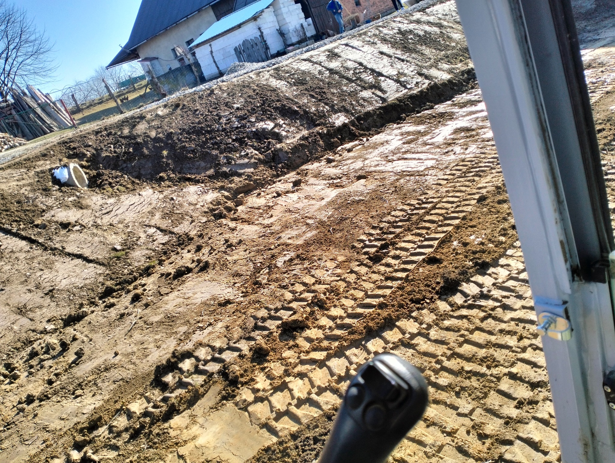 Świeżo wykopany rów w ziemi z widocznymi śladami opon maszyny budowlanej i fragmentem białej rury, widziany z perspektywy operatora maszyny.
