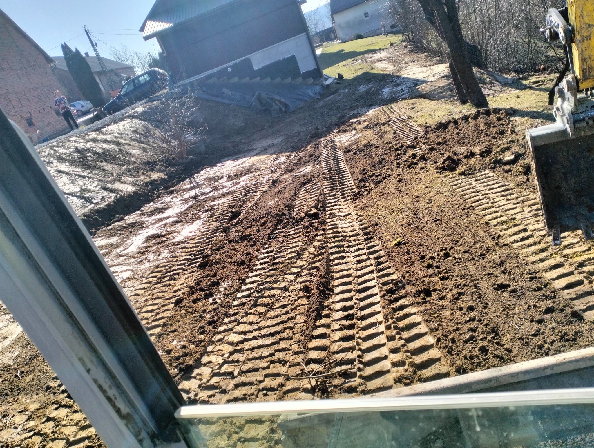 Świeżo wyrównany teren z widocznymi śladami gąsienic koparki, zarys budynku w tle.