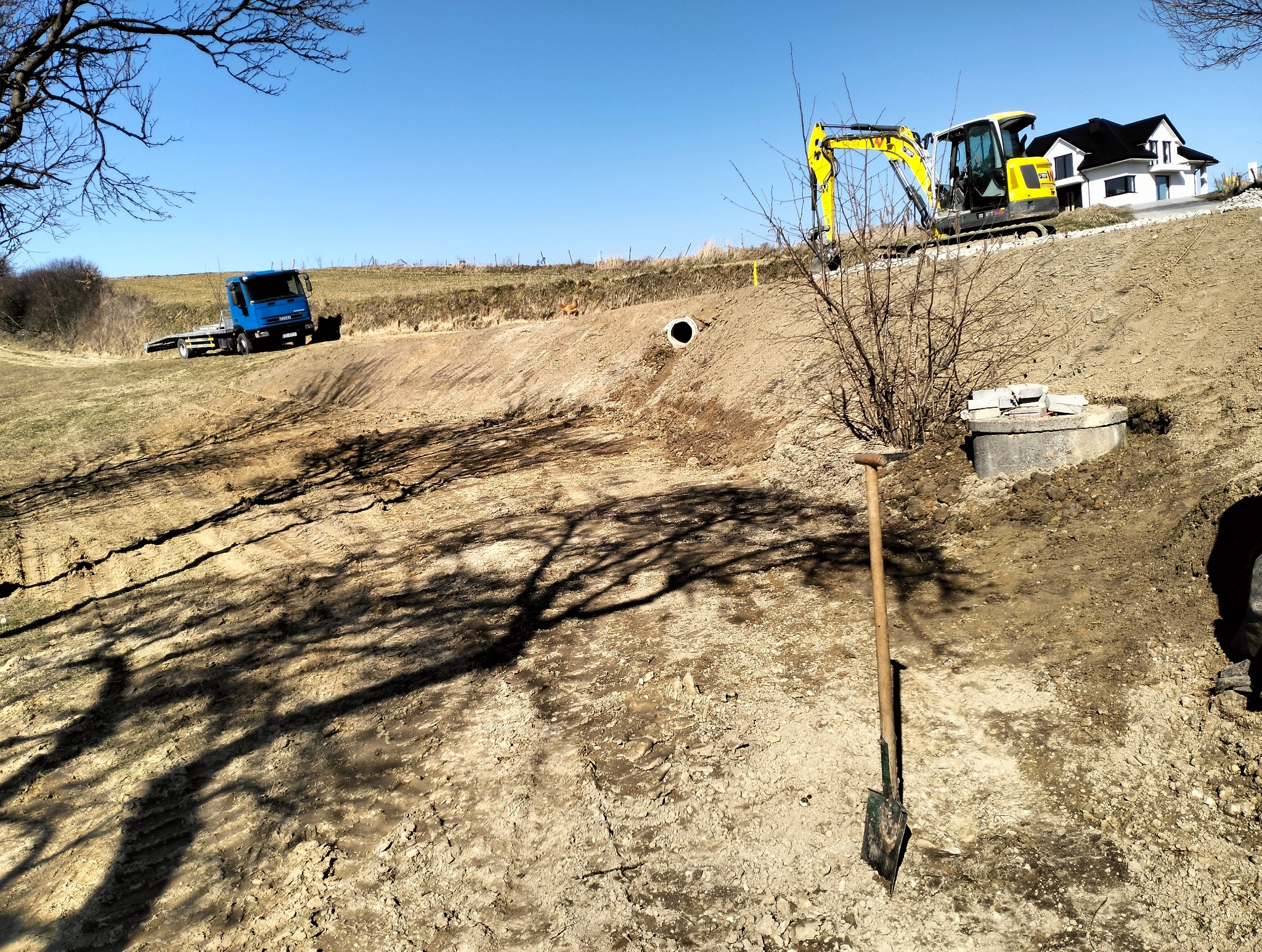 Żółta koparka na zboczu z widocznym wykopem, rurą drenażową i studzienką betonową, w tle niebieska ciężarówka i dom, cień drzewa na pierwszym planie, łopata wbita w ziemię.