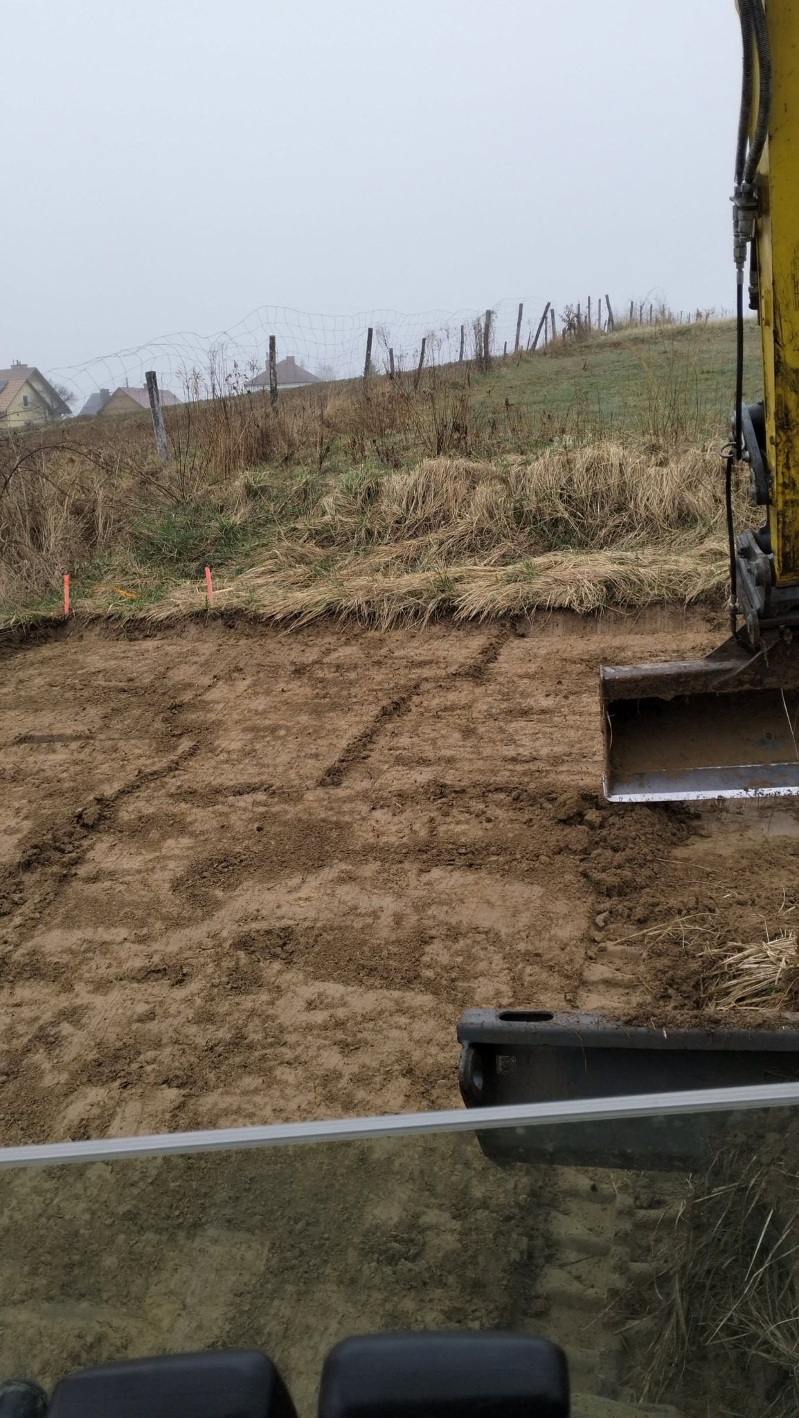 Żółta koparka z łyżką częściowo widoczna, przygotowuje teren pod budowę, widoczne ślady gąsienic na wyrównanej, piaszczystej powierzchni, w tle zarośnięte pole i zabudowania w oddali, pochmurne niebo.