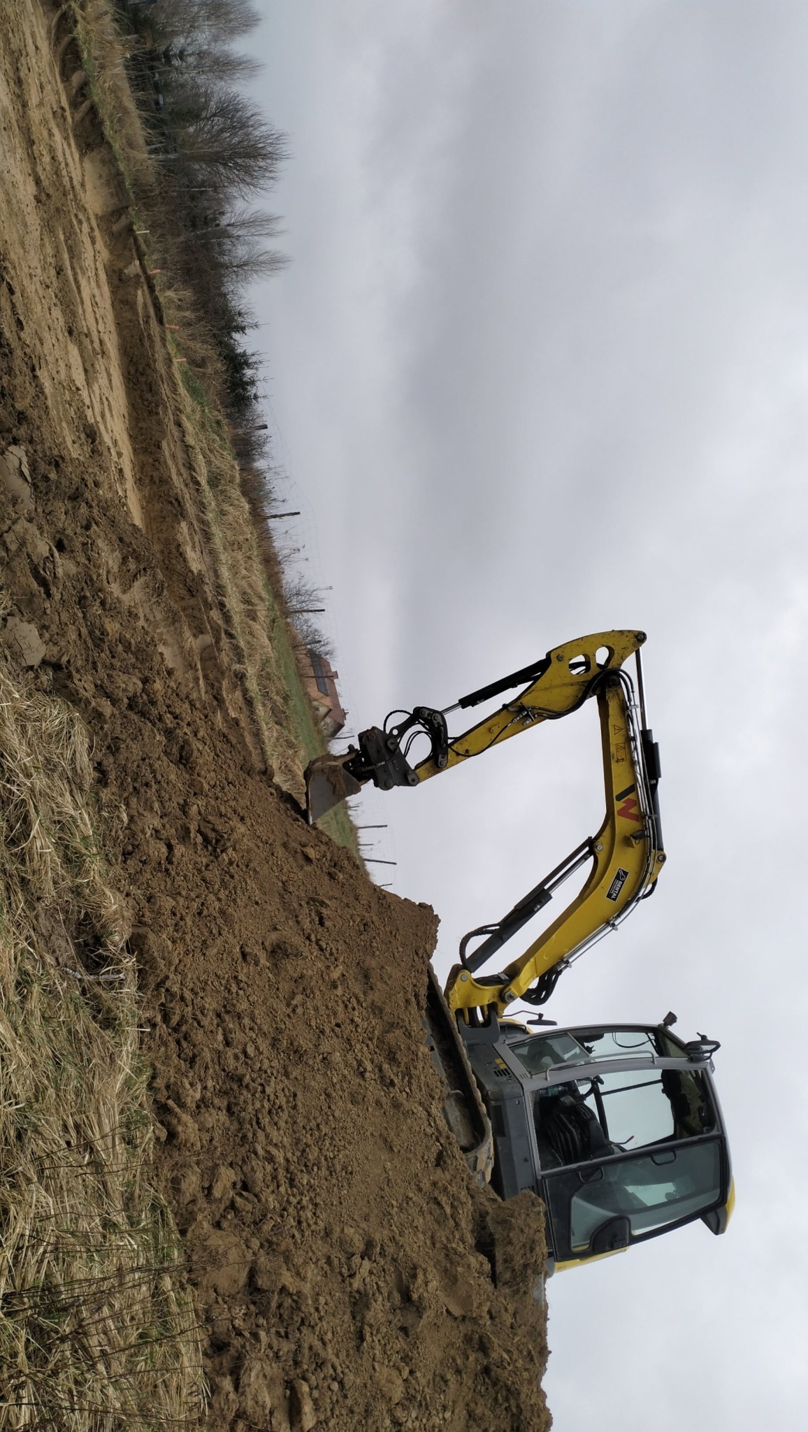 Żółta koparka Wacker Neuson zagłębia łyżkę w ziemi na budowie, widoczne świeże ślady wykopu na tle pochmurnego nieba i odległych zabudowań.