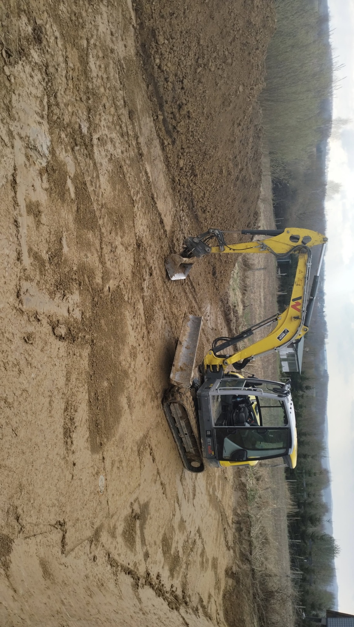 Żółta koparka gąsienicowa na błotnistym terenie, widoczne świeże ślady pracy i pryzma ziemi w tle.