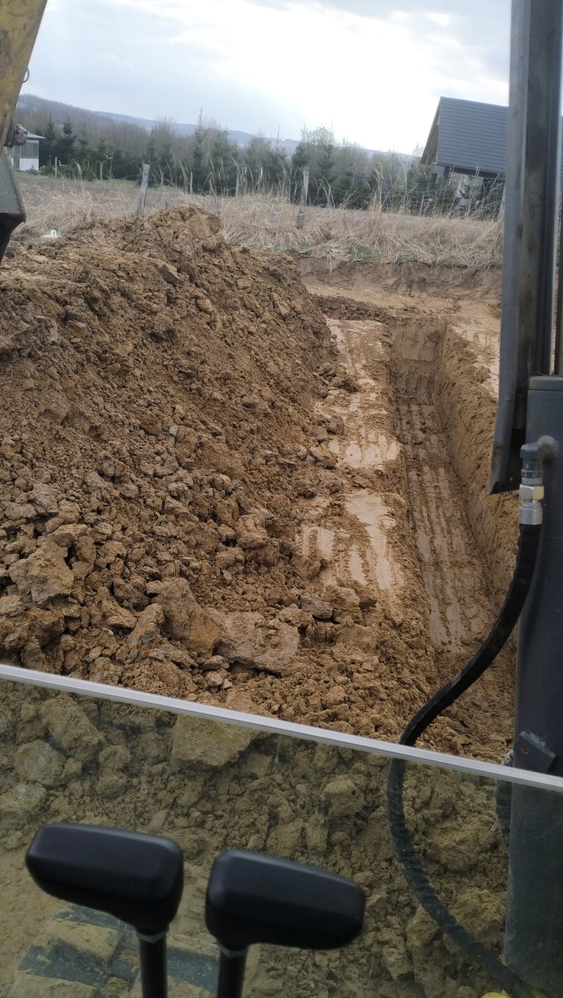 Wykopany rów w ziemi z widocznymi śladami opon, hałda ziemi obok, widok z kabiny maszyny budowlanej na tle drzew i zabudowań.
