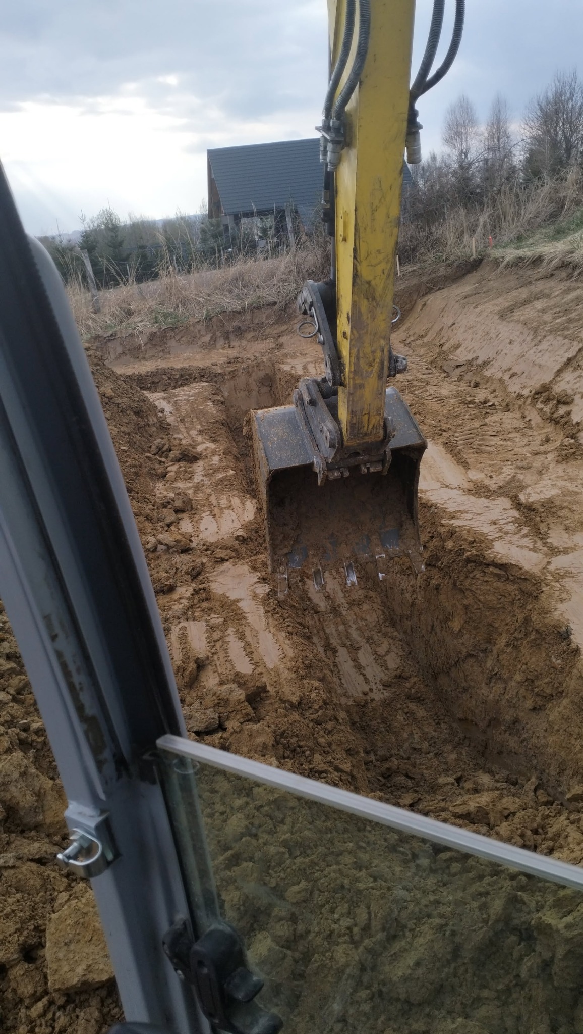 Żółta koparka z łyżką zagłębia się w ziemię, widok z kabiny, w tle dom z ciemnym dachem.