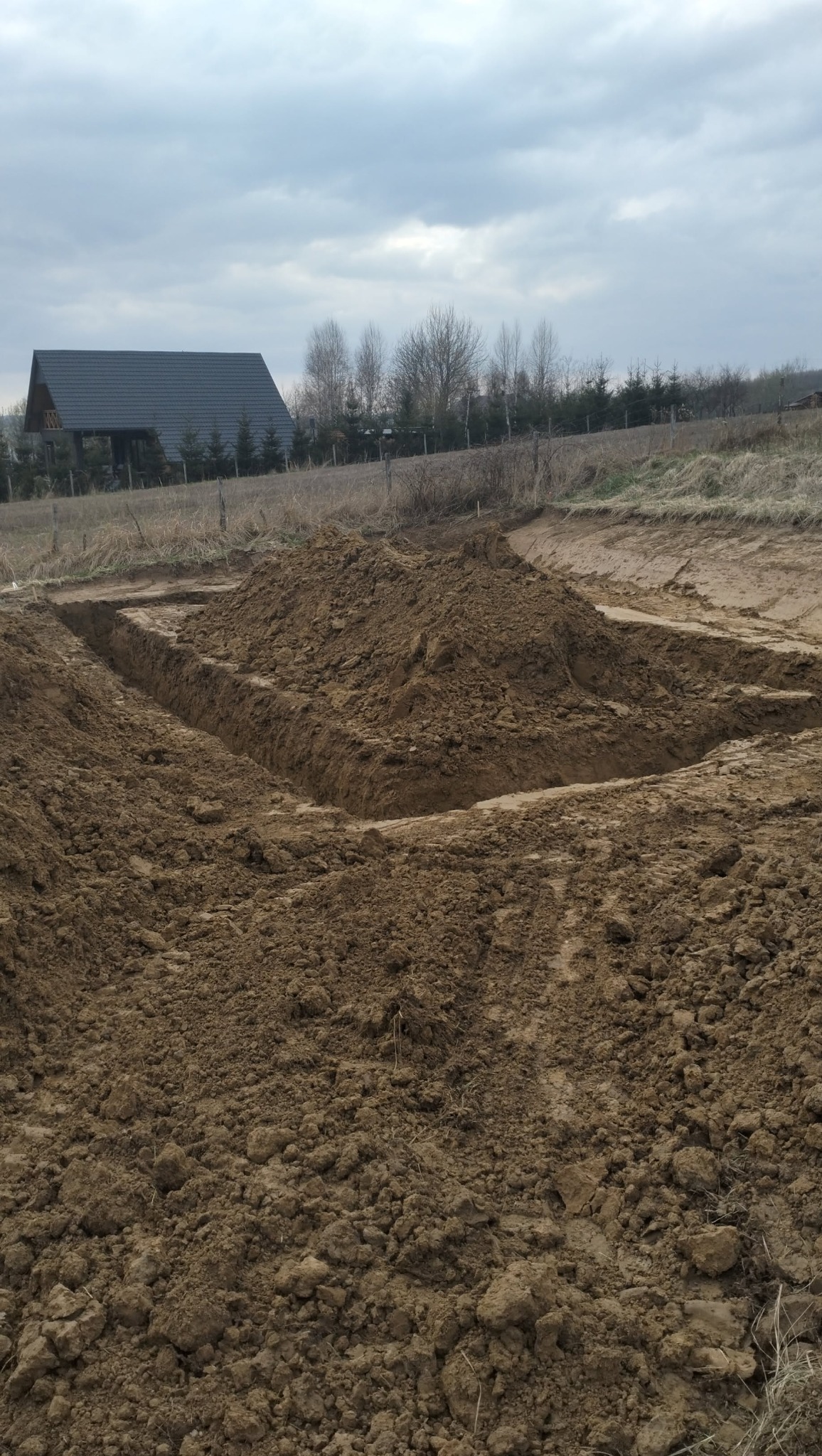 Wykop w kształcie prostokąta, pryzma ziemi wewnątrz, dom z ciemnym dachem w tle, pochmurne niebo.