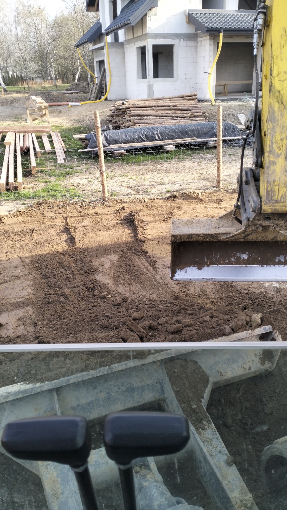 Żółta koparka z łyżką zagłębiającą się w ziemię, widok z kabiny operatora, w tle budynek w stanie surowym otoczony ogrodzeniem z siatki i stertą drewna.