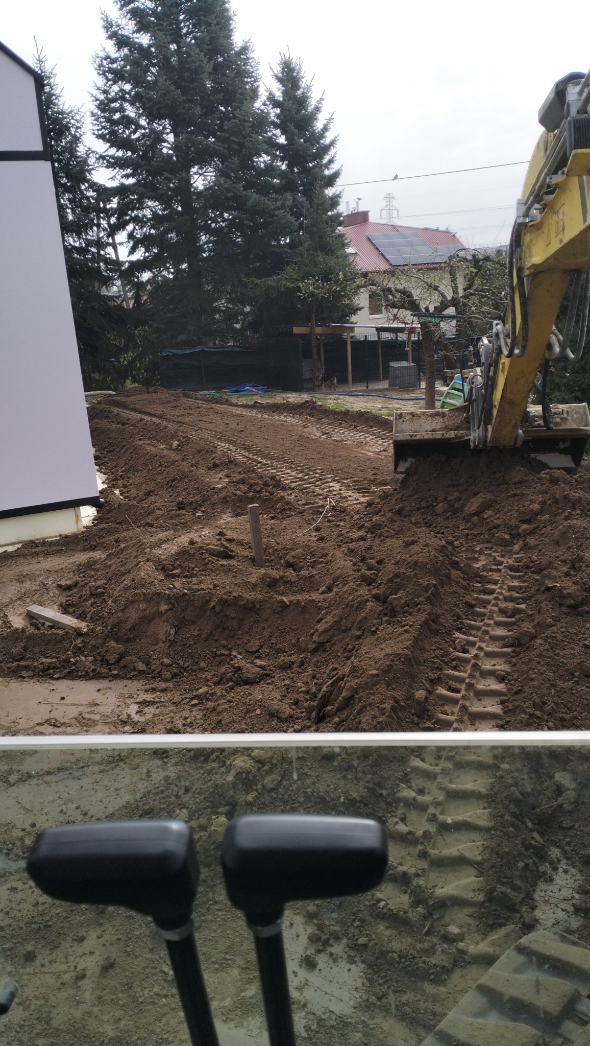 Żółta koparka w trakcie prac ziemnych, widoczne świeże ślady gąsienic na wyrównanym terenie, w tle dom z panelami słonecznymi i wysokie drzewa iglaste. Widok z kabiny operatora.