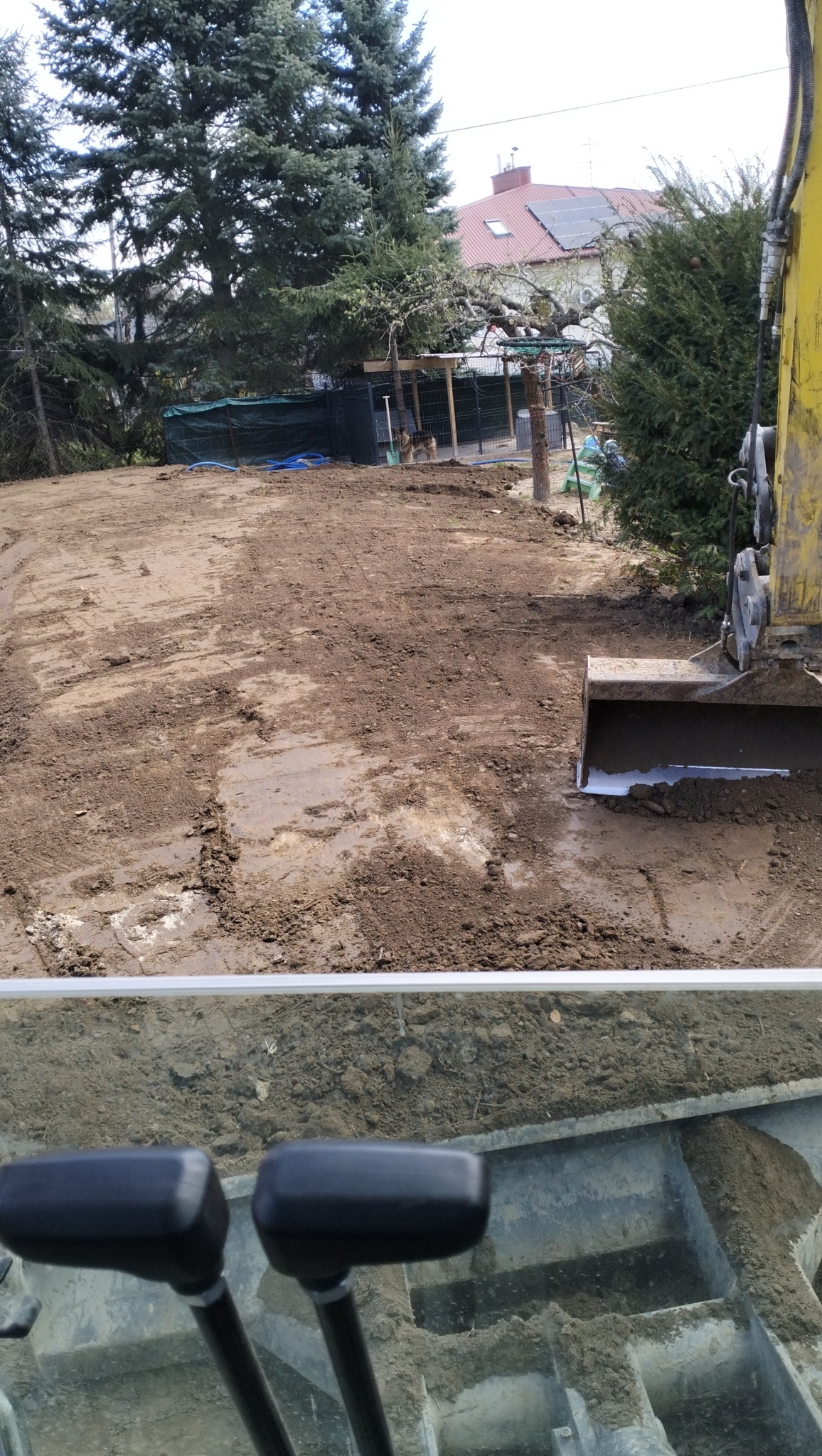 Widok z kabiny koparki na teren w trakcie wyrównywania, z widocznymi śladami opon i łyżką koparki w tle, drzewa i budynek w oddali.