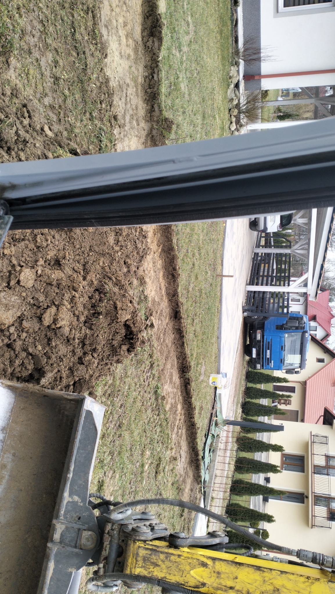 Wykop w ziemi, widok z kabiny koparki, w tle dom z czerwonym dachem i niebieska ciężarówka.