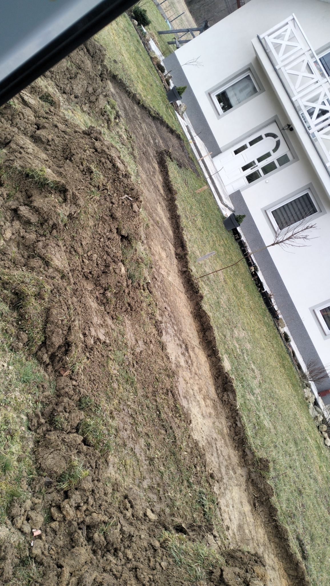 Wykopany wąski rów w trawniku wzdłuż ściany domu, widoczne świeże ślady ziemi i wykopu.