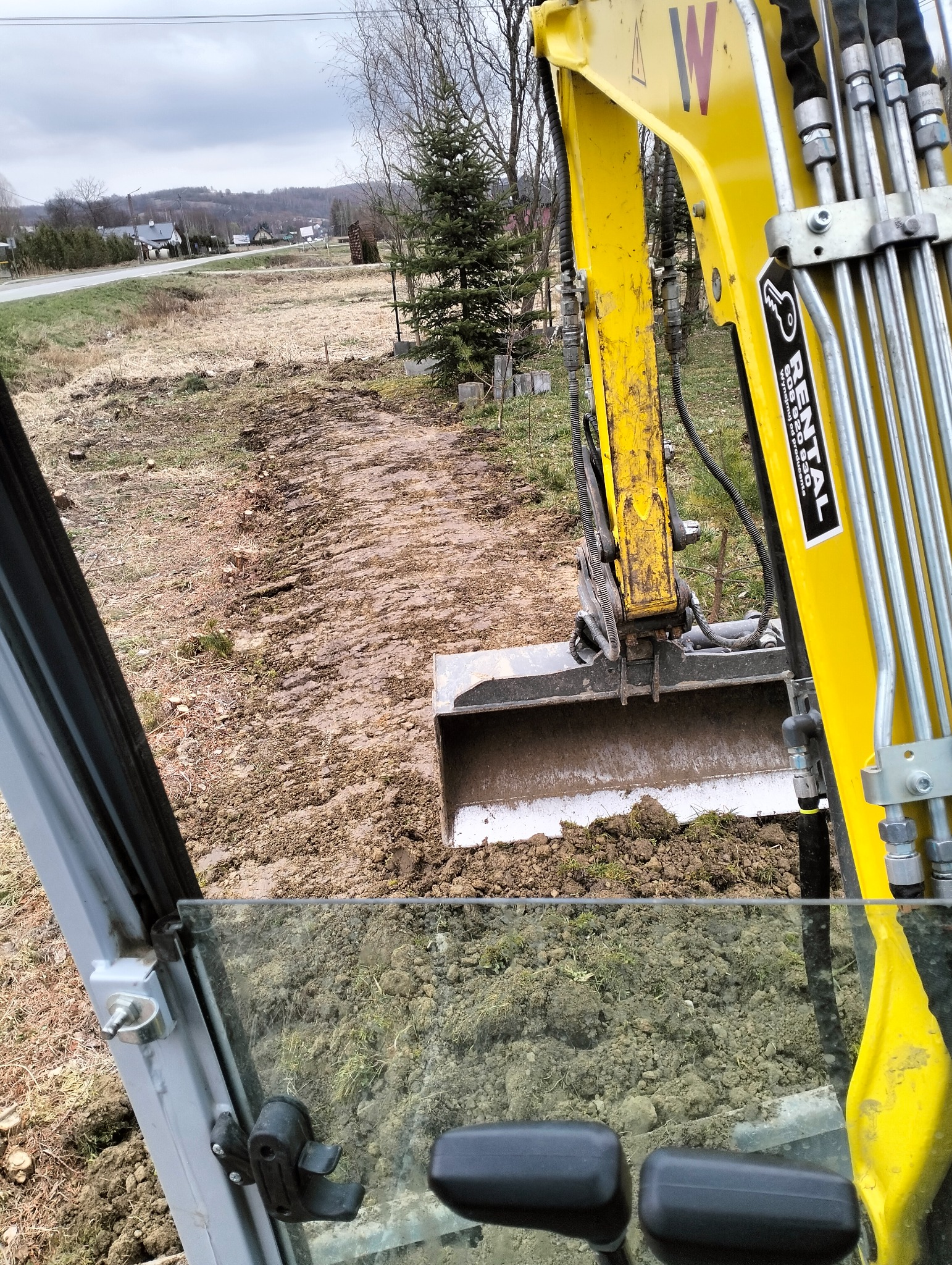 Widok z kabiny koparki na wykopany pas ziemi na działce z widocznymi domami w tle, brudna łyżka koparki wypełniona ziemią. Na ramieniu koparki naklejka firmy wypożyczającej sprzęt budowlany.