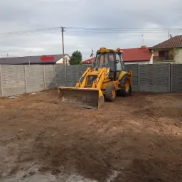 Żółta koparko-ładowarka na placu budowy, w tle szare panele ogrodzeniowe o fakturze betonu i budynki mieszkalne pod pochmurnym niebem.