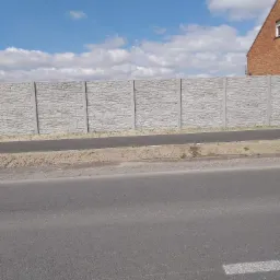 Długi, szary mur z prefabrykatów o fakturze kamienia, oddzielający drogę od terenu posesji, z fragmentem ceglanego budynku w tle i błękitnym niebem z obłokami.