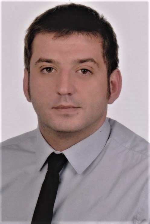 Portret mężczyzny w szarej koszuli i czarnym krawacie, zdjęcie paszportowe z delikatnym uśmiechem.