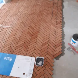 Montaż ceglastej terakoty w jodełkę, widoczny pojemnik z krzyżykami dystansowymi, puszka z klejem i narzędzia, częściowo ułożona podłoga.