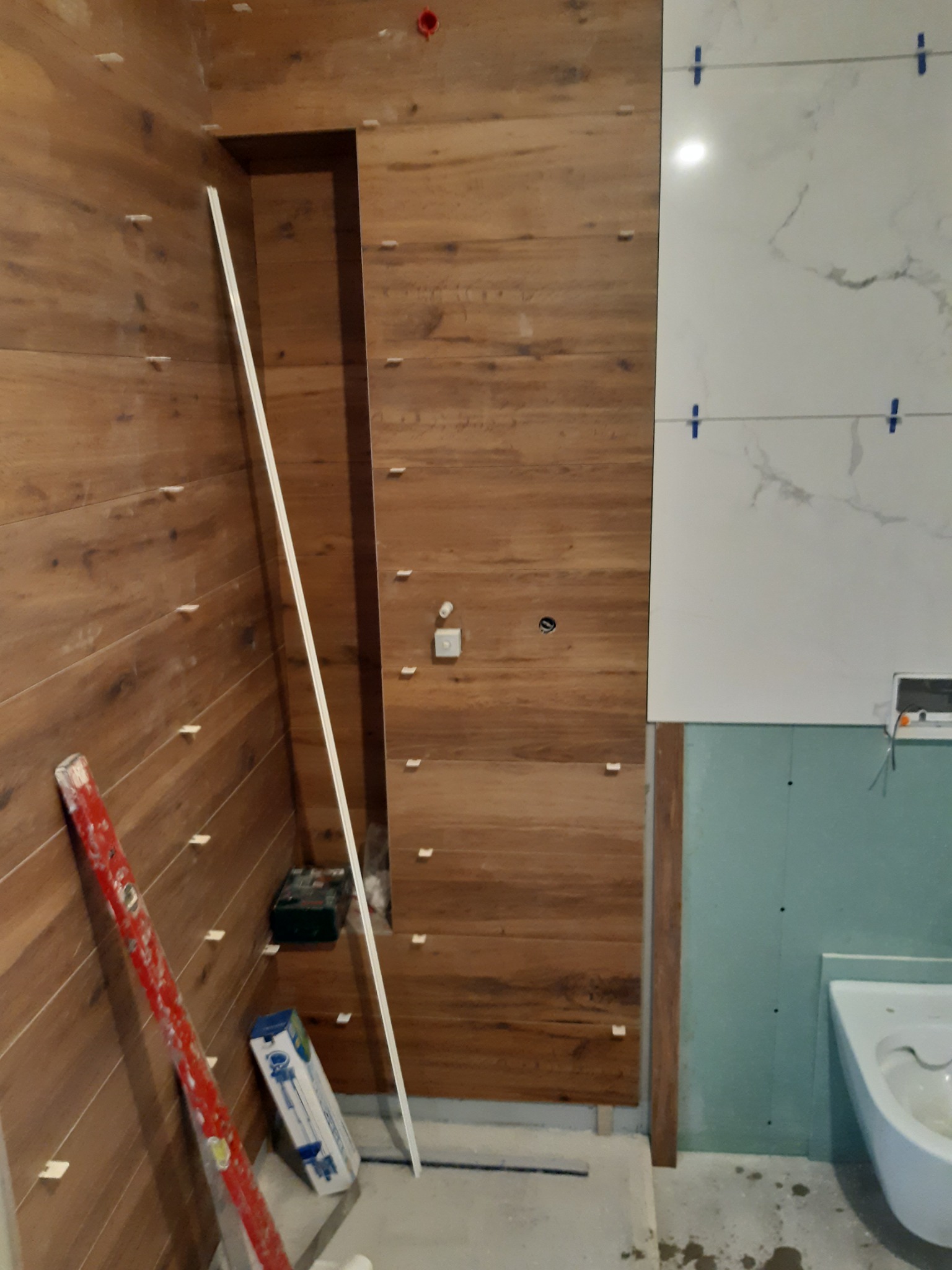 Montaż płytek imitujących drewno na ścianie z wnęką obok częściowo wykończonej ściany z białych płytek marmurkowych, widoczne krzyżyki dystansowe, poziomica i narzędzia.