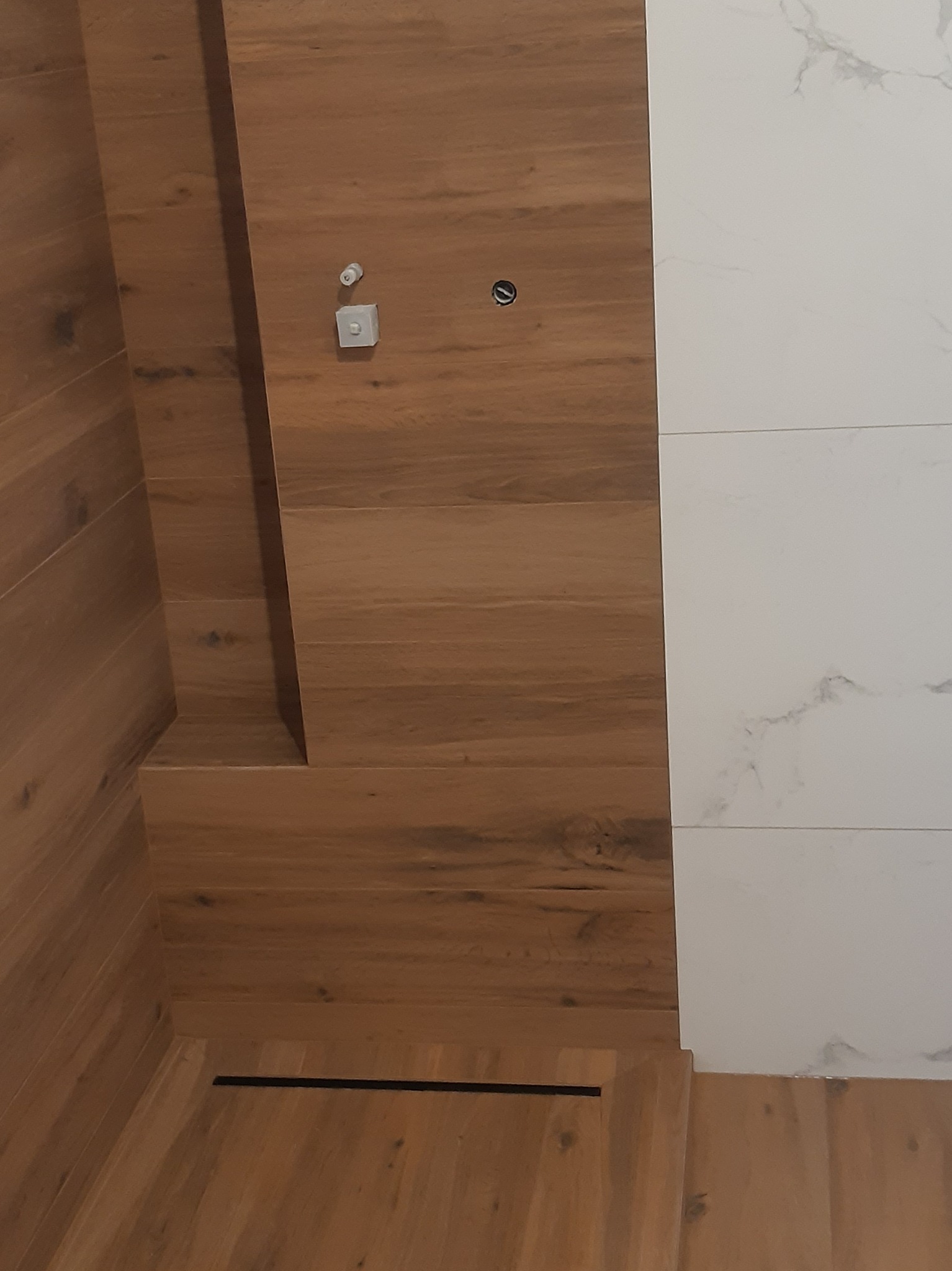 Fragment łazienki z wykończeniem ścian w drewnie i białych płytkach imitujących marmur, widoczny odpływ liniowy w podłodze i elementy instalacji.