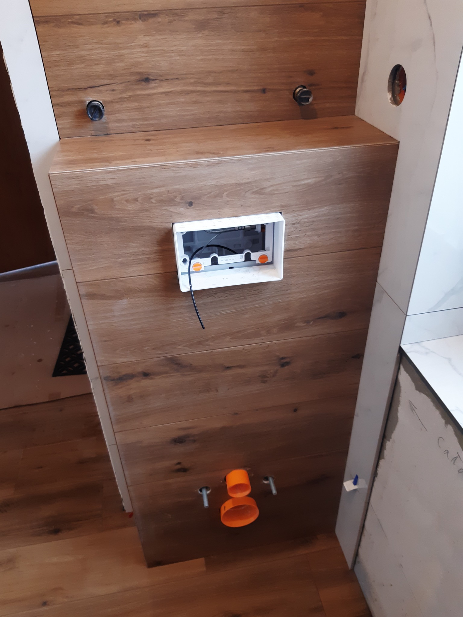 Ścianka zabudowy stelaża WC wykończona panelami drewnopodobnymi, widoczna instalacja wodno-kanalizacyjna oraz puszka elektryczna pod przycisk spłukujący.