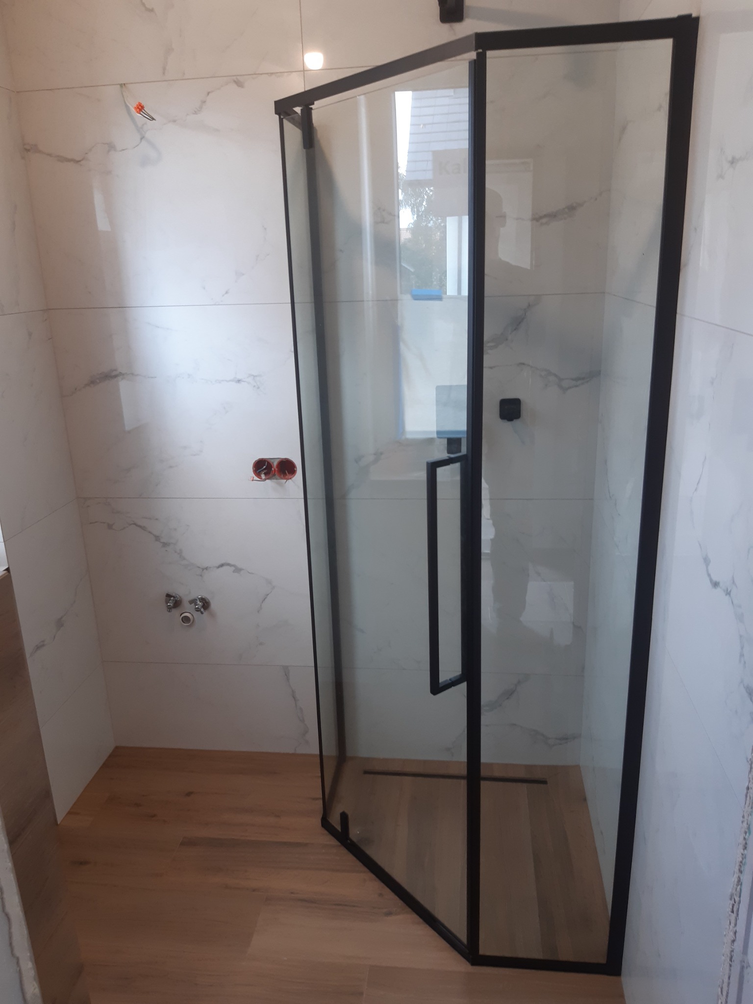 Kabina prysznicowa z czarnymi profilami, zamontowana w łazience z białymi płytkami imitującymi marmur i drewnopodobną podłogą, widoczne niezamaskowane gniazdka elektryczne i przyłącze wodne.