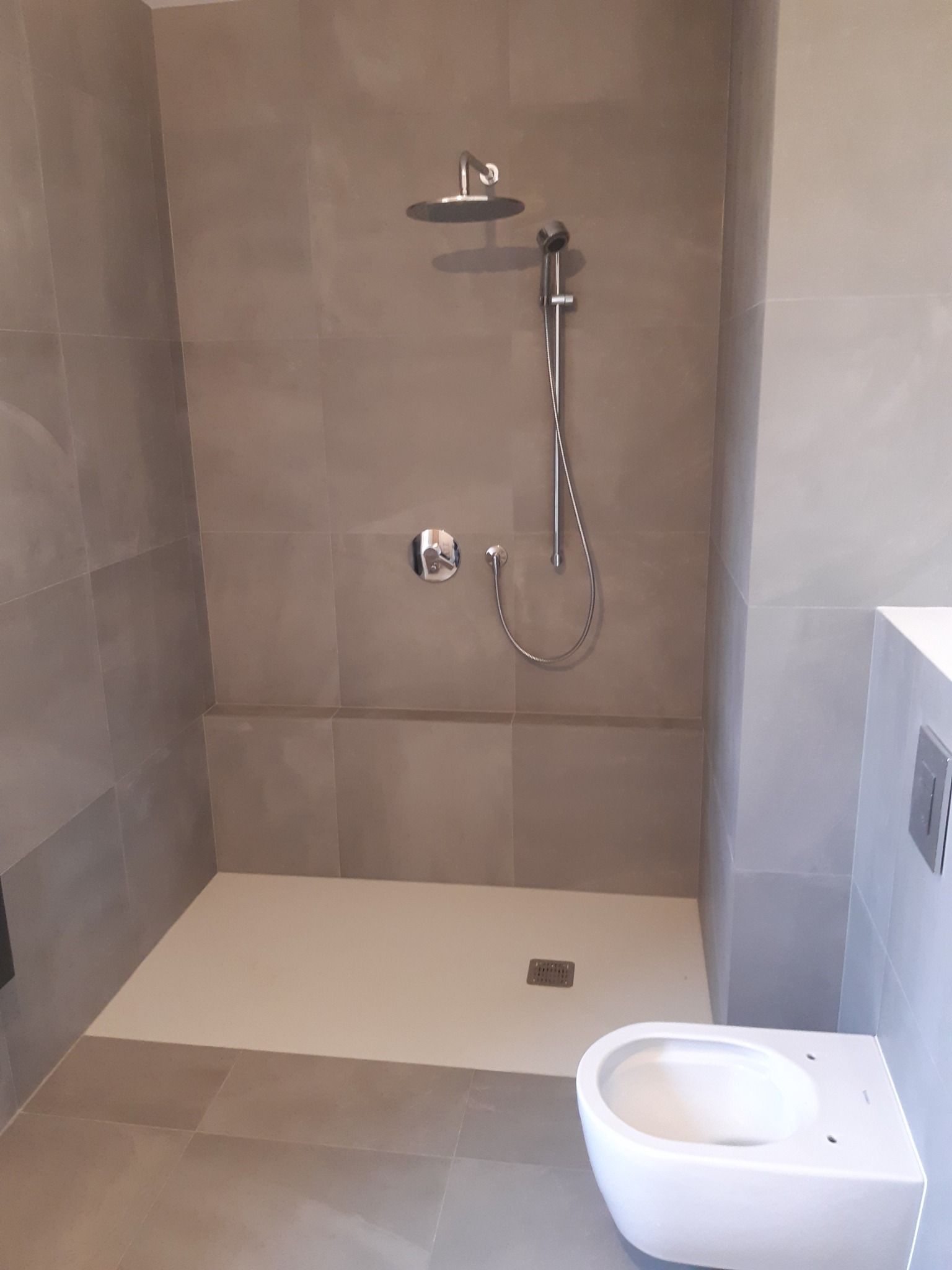 Nowoczesna kabina prysznicowa z deszczownicą i panelem prysznicowym, wykończona szarą płytką wielkoformatową, z widoczną miską WC w minimalistycznym stylu.