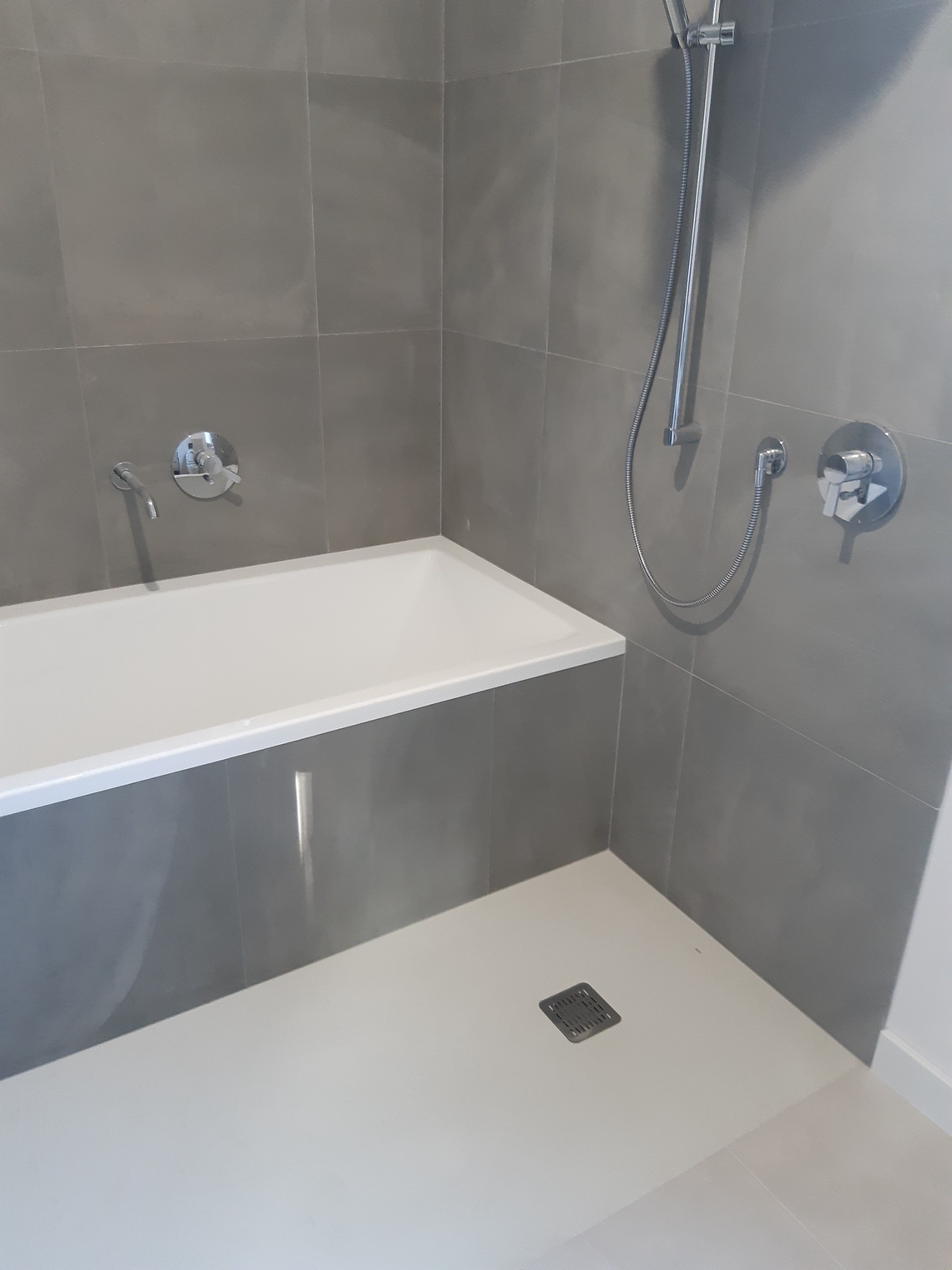 Nowoczesna łazienka z szarą glazurą, białą wanną i kabiną prysznicową z chromowaną armaturą, widoczny odpływ liniowy.