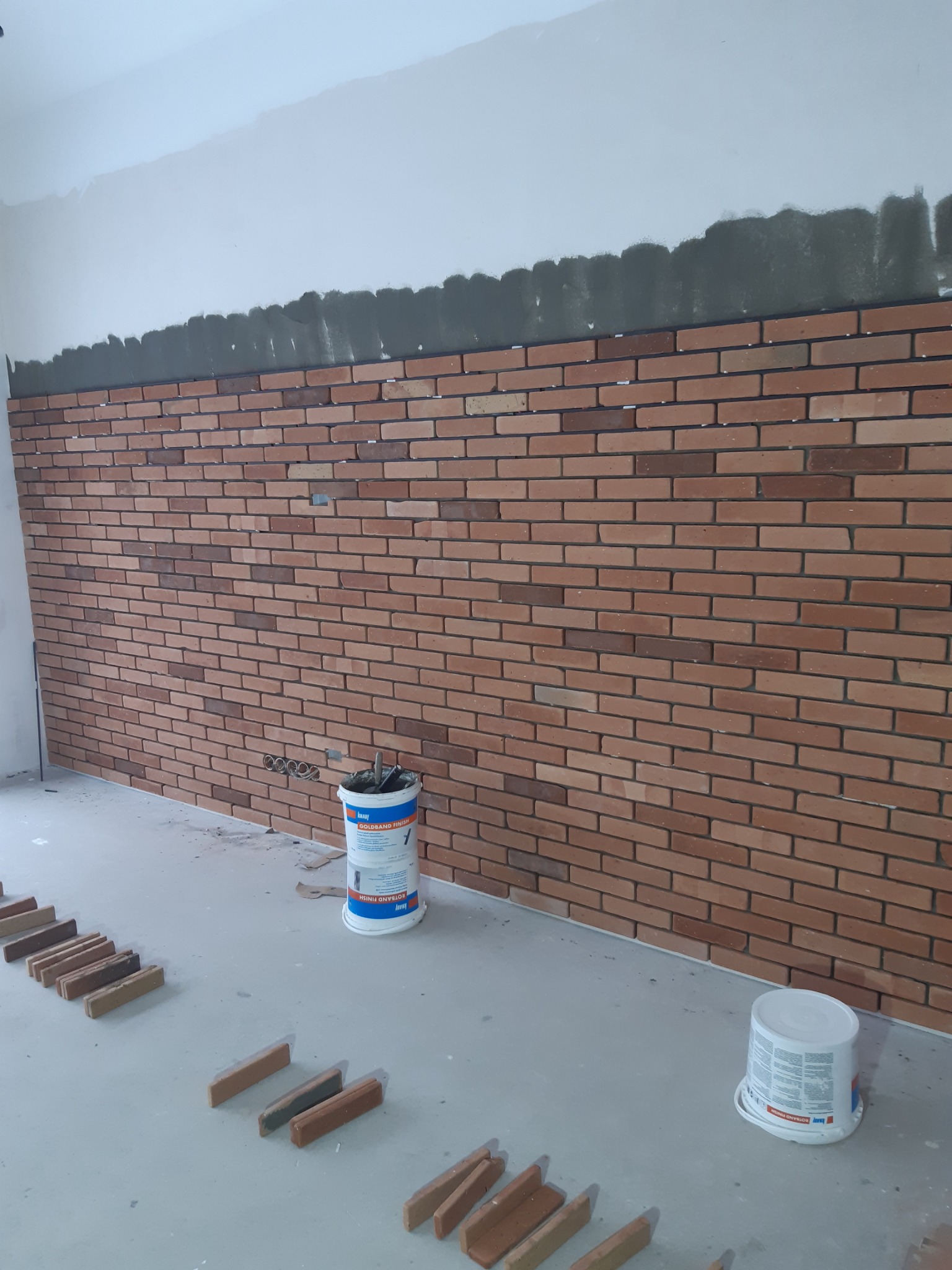Ściana wyłożona cegiełkami klinkierowymi w trakcie montażu, widoczne wiaderka z zaprawą Knauf Goldband Finish i Rotband Finish oraz ułożone na podłodze cegiełki przygotowane do przyklejenia.