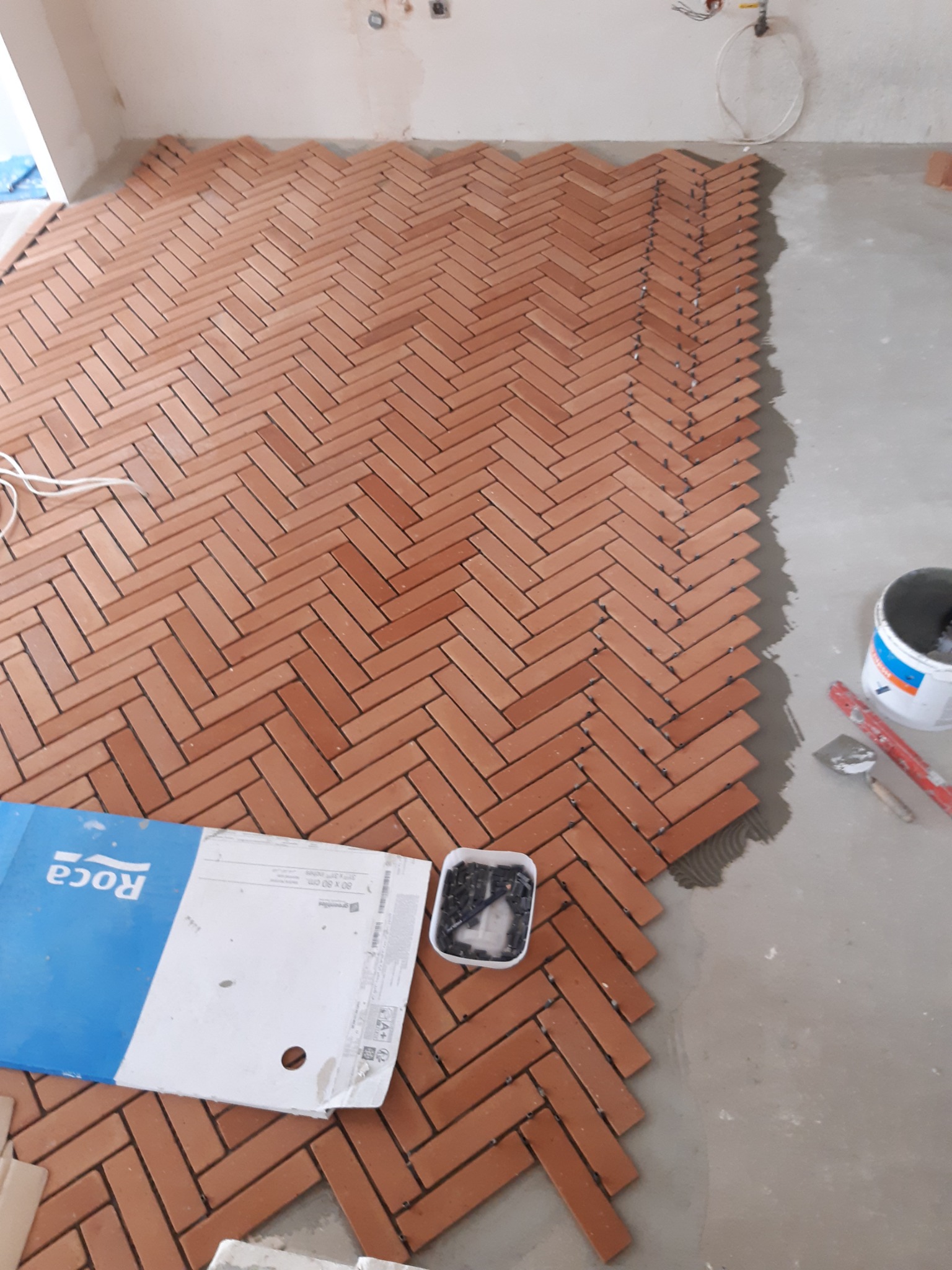 Montaż ceglastej terakoty w jodełkę, widoczny pojemnik z krzyżykami dystansowymi, puszka z klejem i narzędzia, częściowo ułożona podłoga.