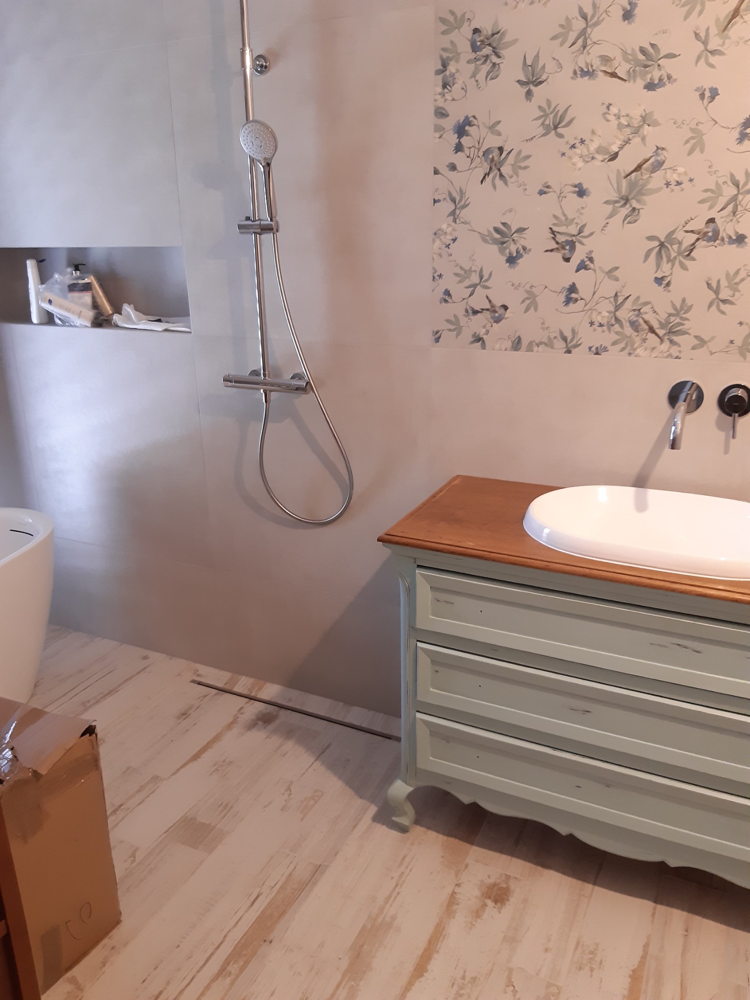 Umywalka nablatowa na odrestaurowanej komodzie w łazience z kwiatową tapetą, chromowana bateria ścienna i zestaw prysznicowy.