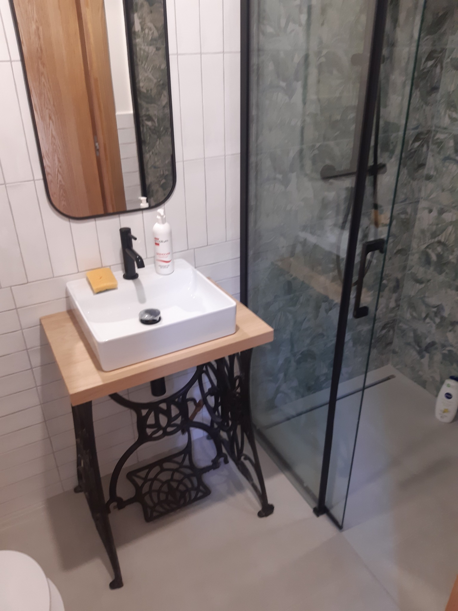 Biała umywalka nablatowa z czarną armaturą, osadzona na drewnianym blacie wspartym na odrestaurowanej podstawie maszyny do szycia Singer, kabina prysznicowa z wzorem liści w tle, białe płytki...