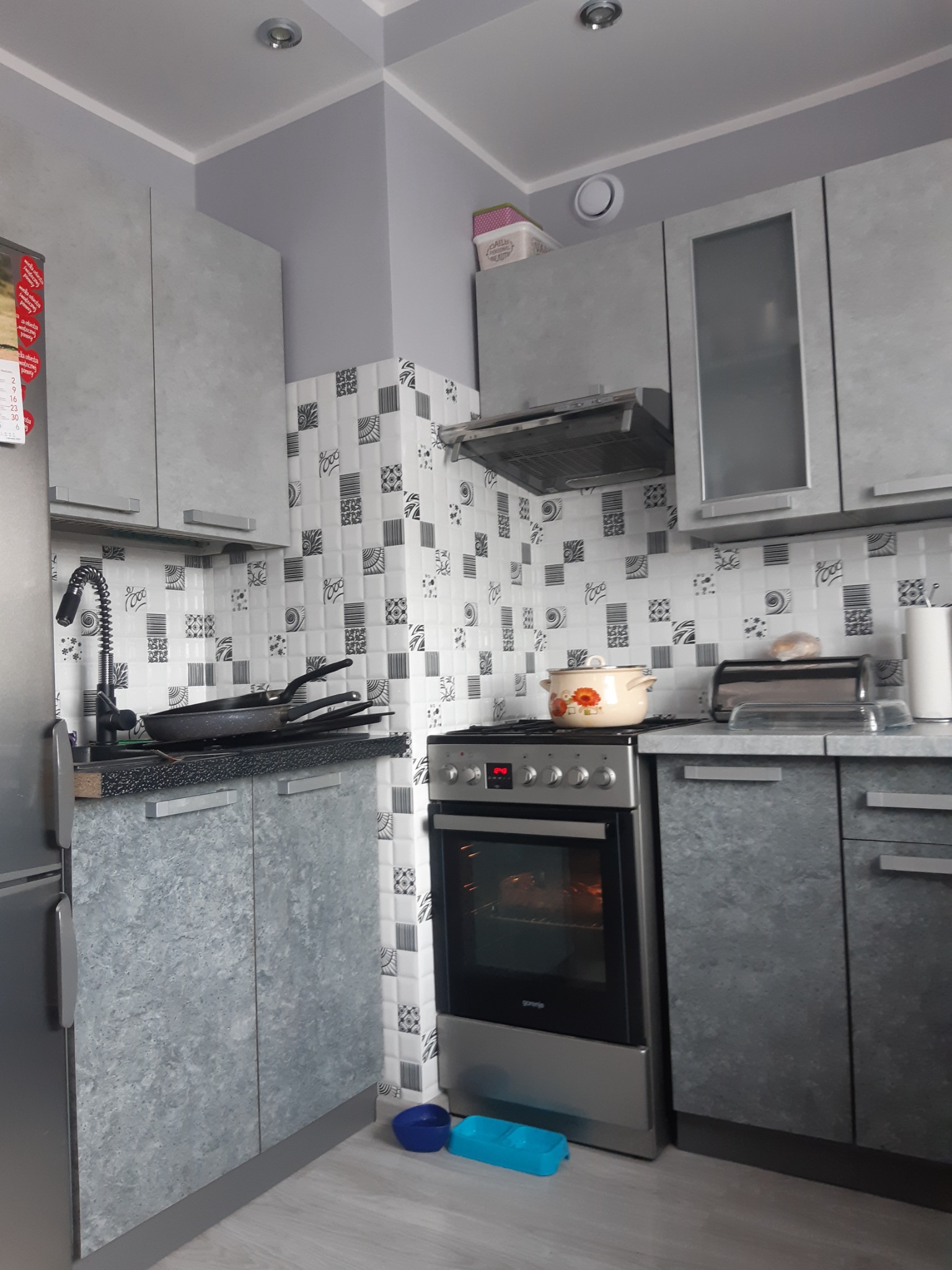 Szara kuchnia z wzorzystymi kaflami, garnkiem na kuchence i stosem patelni przy zlewie.