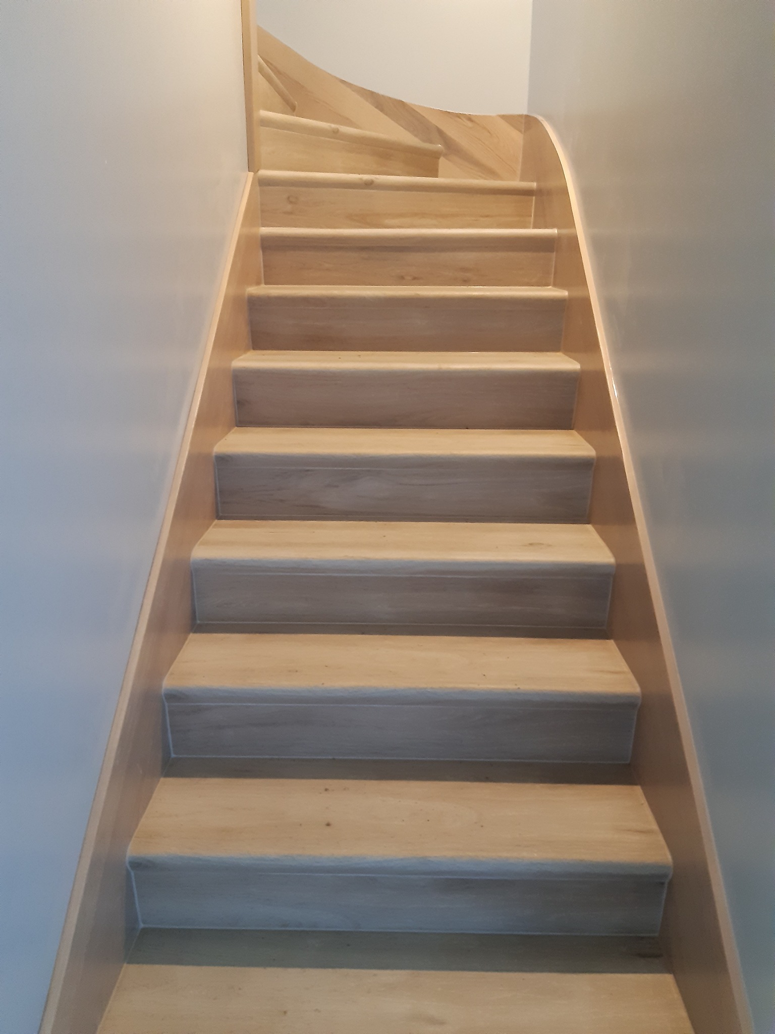 Jasne drewniane schody z zaokrąglonymi krawędziami stopni, prowadzące w górę, oświetlone z góry, przylegające do pomalowanych na szaro ścian.