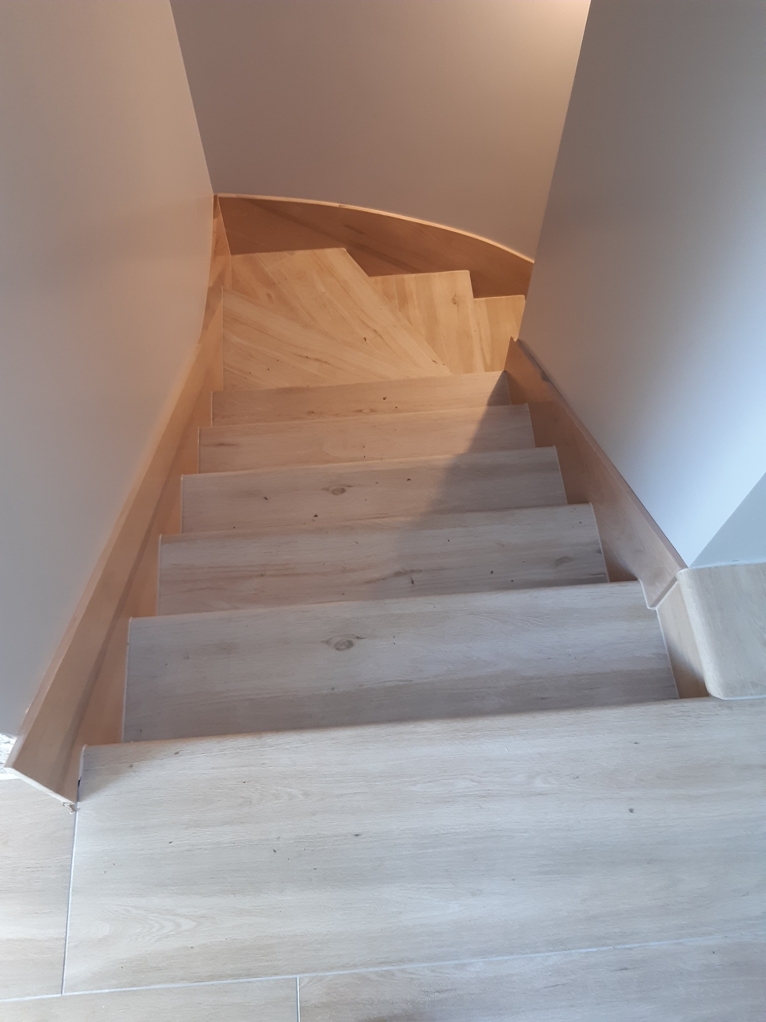 Drewniane schody z delikatnym usłojeniem, prowadzące w dół, z widocznymi stopniami i listwami przypodłogowymi, w jasnym wnętrzu.