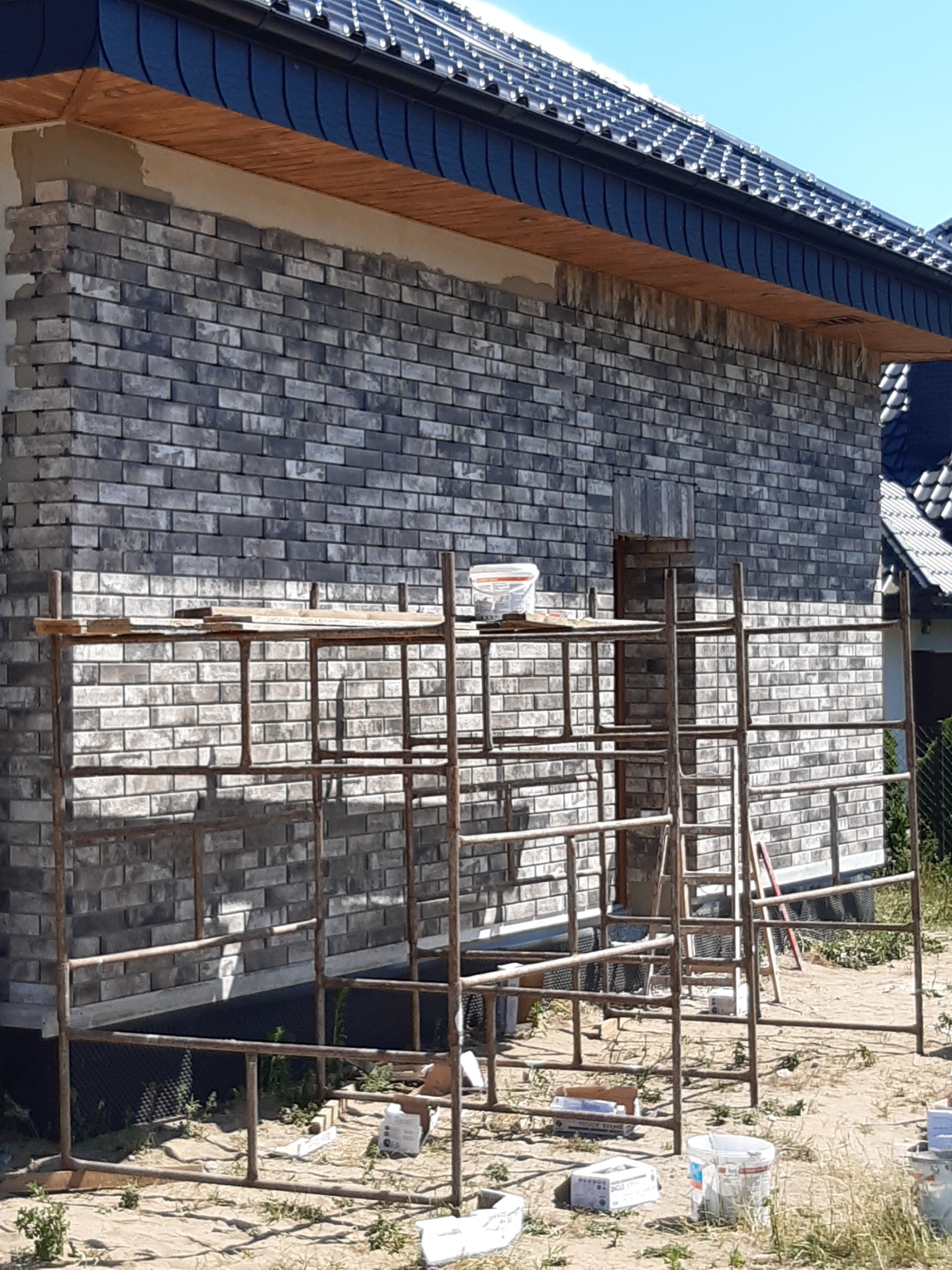 Ściana domu w trakcie wykańczania klinkierem w odcieniach szarości, widoczne rusztowanie, wiaderko z zaprawą na górze rusztowania, nieukończone fragmenty elewacji.
