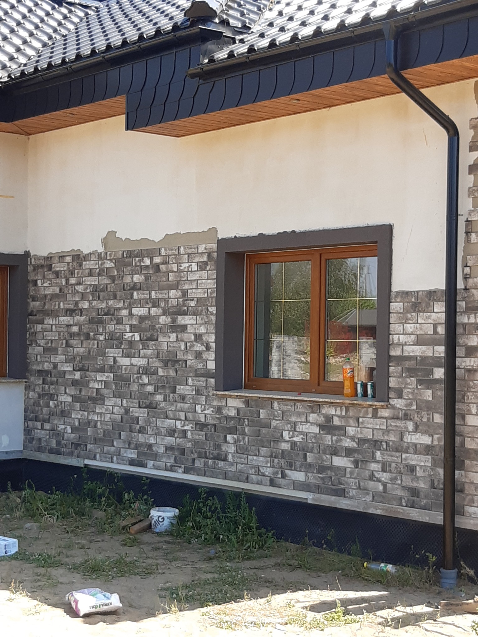 Ściana budynku w trakcie wykończenia z szarej cegły klinkierowej, widoczne okno z brązową ramą i szarym obramowaniem, butelka i puszki na parapecie, częściowo otynkowana ściana powyżej, czarna...