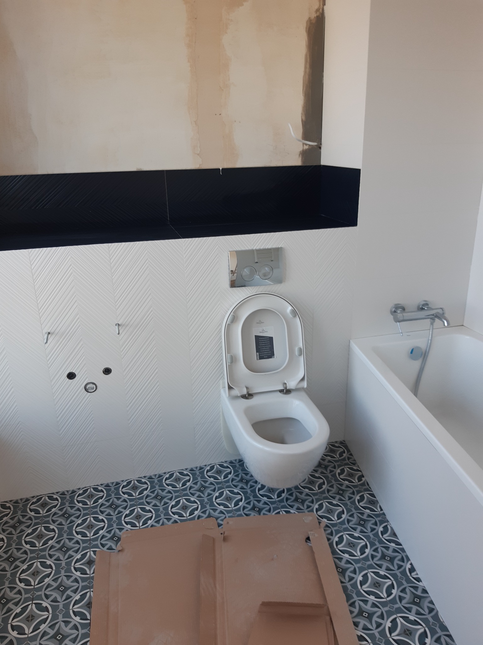 Wnętrze łazienki w trakcie remontu: białe płytki w jodełkę na ścianach, wzorzysta szaro-niebieska podłoga, biała toaleta z otwartą klapą i fragment wanny, widoczne niedokończone prace wykończeniowe...