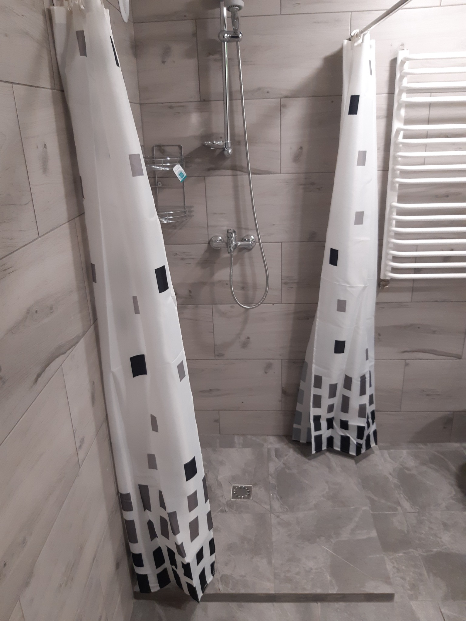Łazienka z szarymi płytkami imitującymi drewno na ścianach i podłodze, kabina prysznicowa z białą zasłoną w czarne i szare kwadraty, chromowana bateria i półka prysznicowa, grzejnik drabinkowy.
