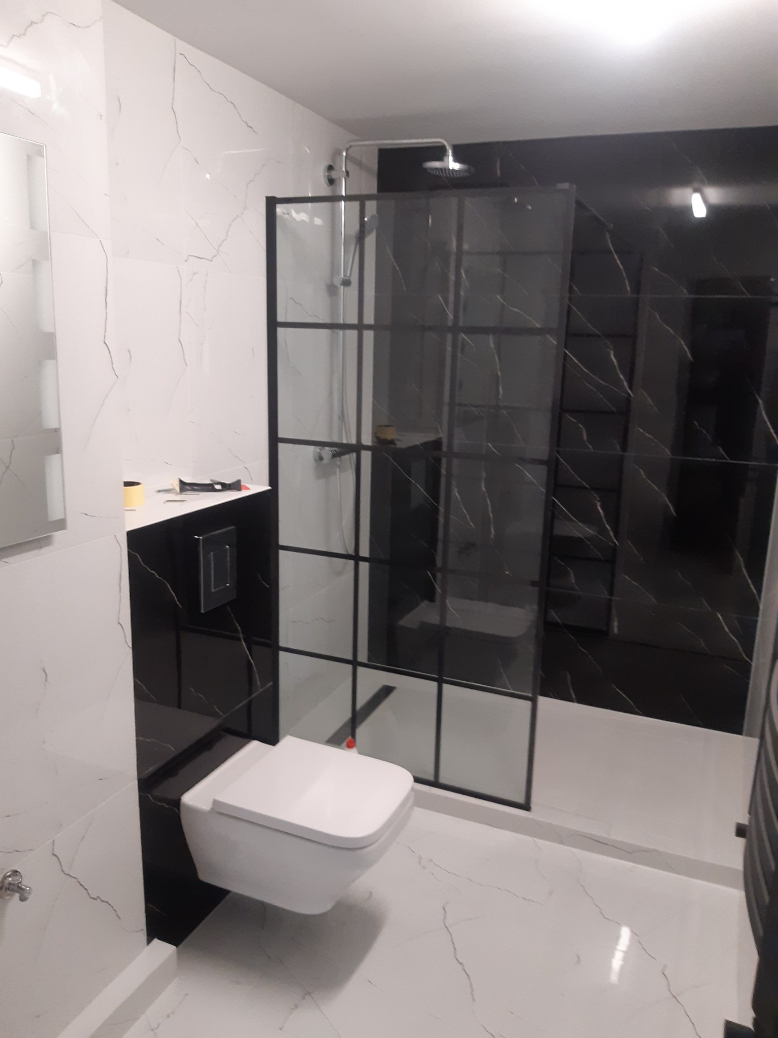 Nowoczesna łazienka z białymi i czarnymi marmurowymi płytkami, kabiną prysznicową typu walk-in z czarnymi szprosami i podwieszaną toaletą.