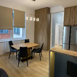 Jasne wnętrze mieszkania po remoncie: stół z czarnymi krzesłami, drewniane panele podłogowe, zabudowa kuchenna z lodówką i oknami z beżowymi roletami rzymskimi.