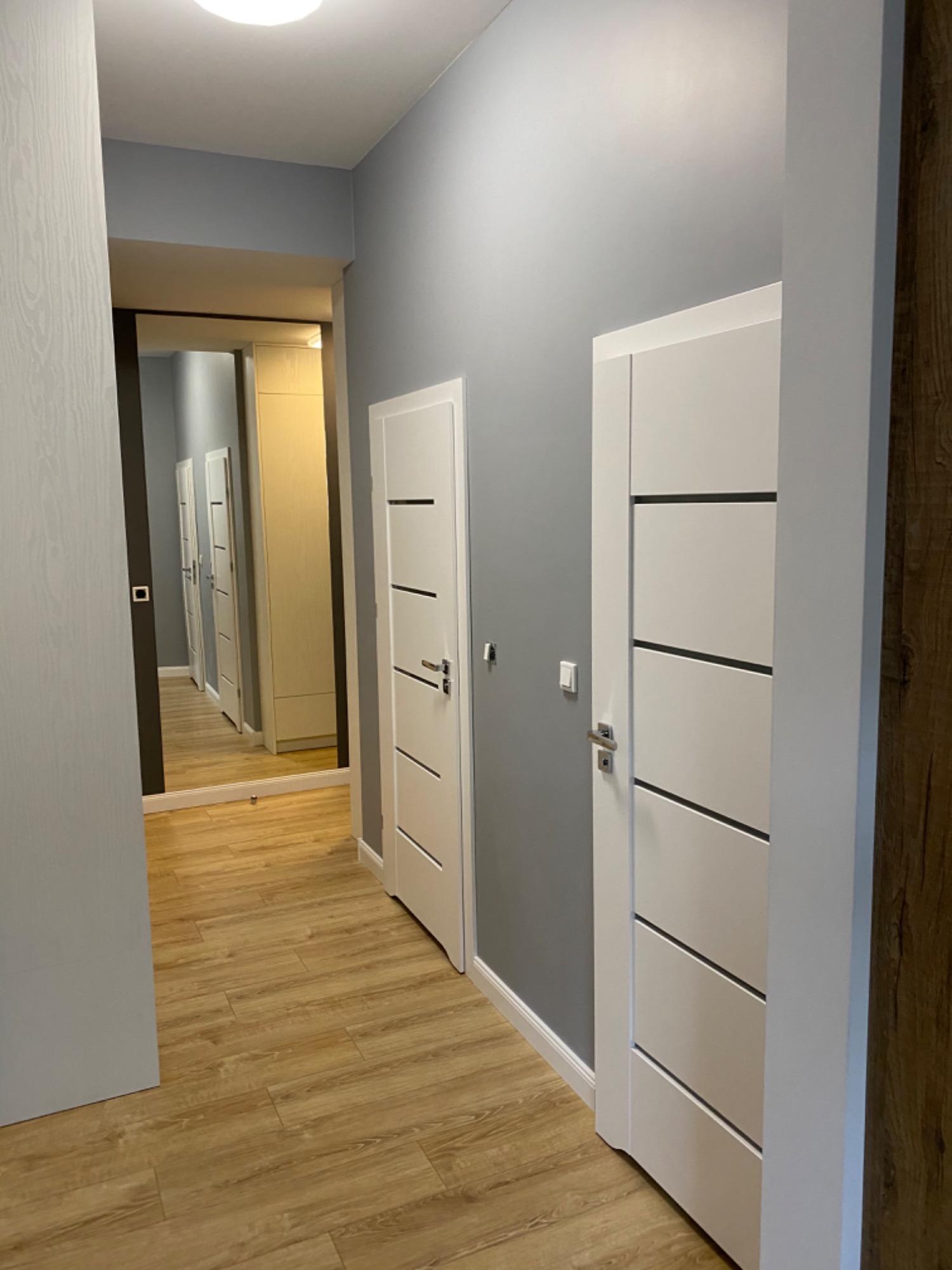 Korytarz w odcieniach szarości z drewnianą podłogą, białymi drzwiami z poziomymi frezami i lustrzanymi drzwiami szafy w tle, odbijającymi dalszą część korytarza.