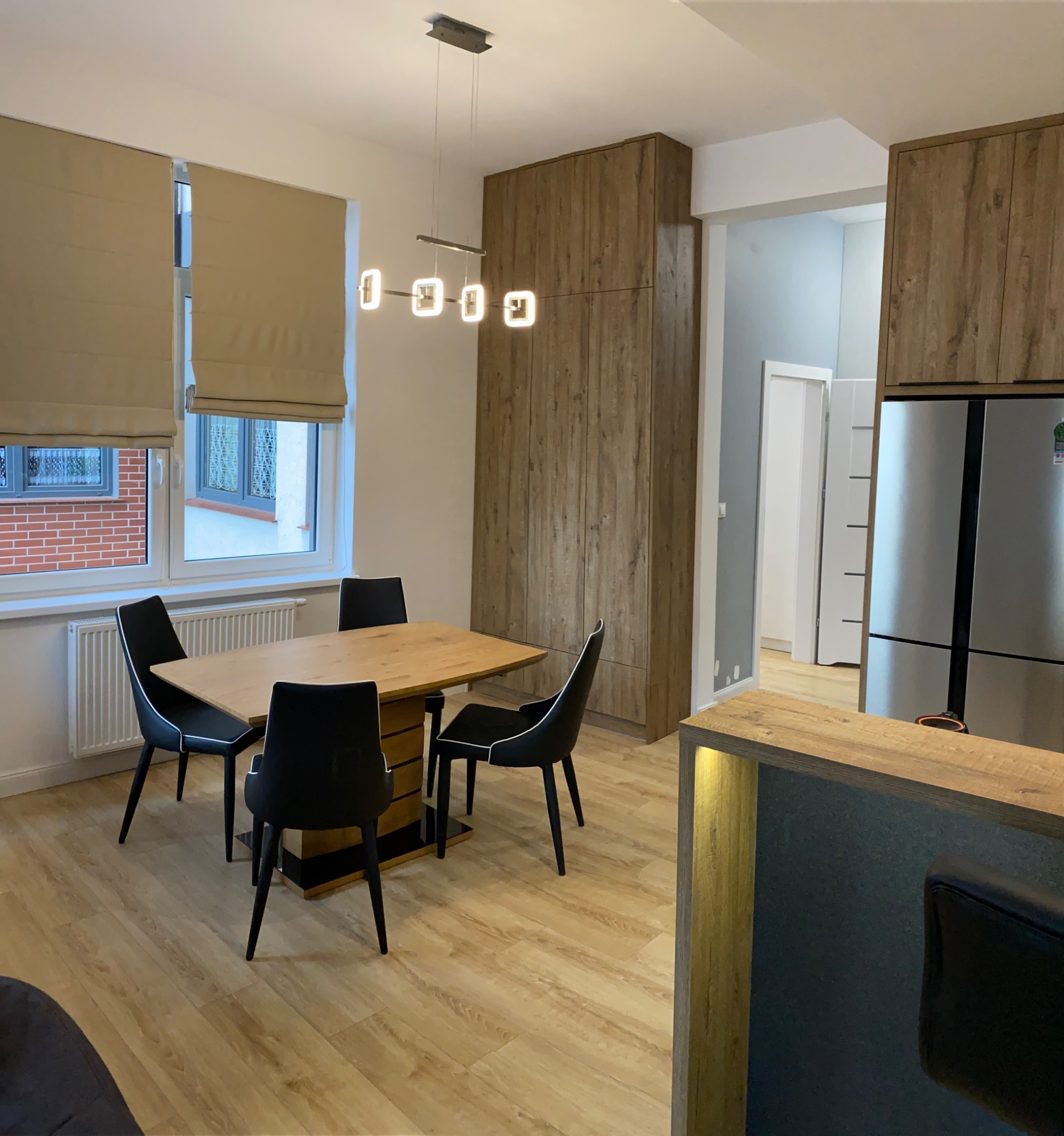 Jasne wnętrze mieszkania po remoncie: stół z czarnymi krzesłami, drewniane panele podłogowe, zabudowa kuchenna z lodówką i oknami z beżowymi roletami rzymskimi.