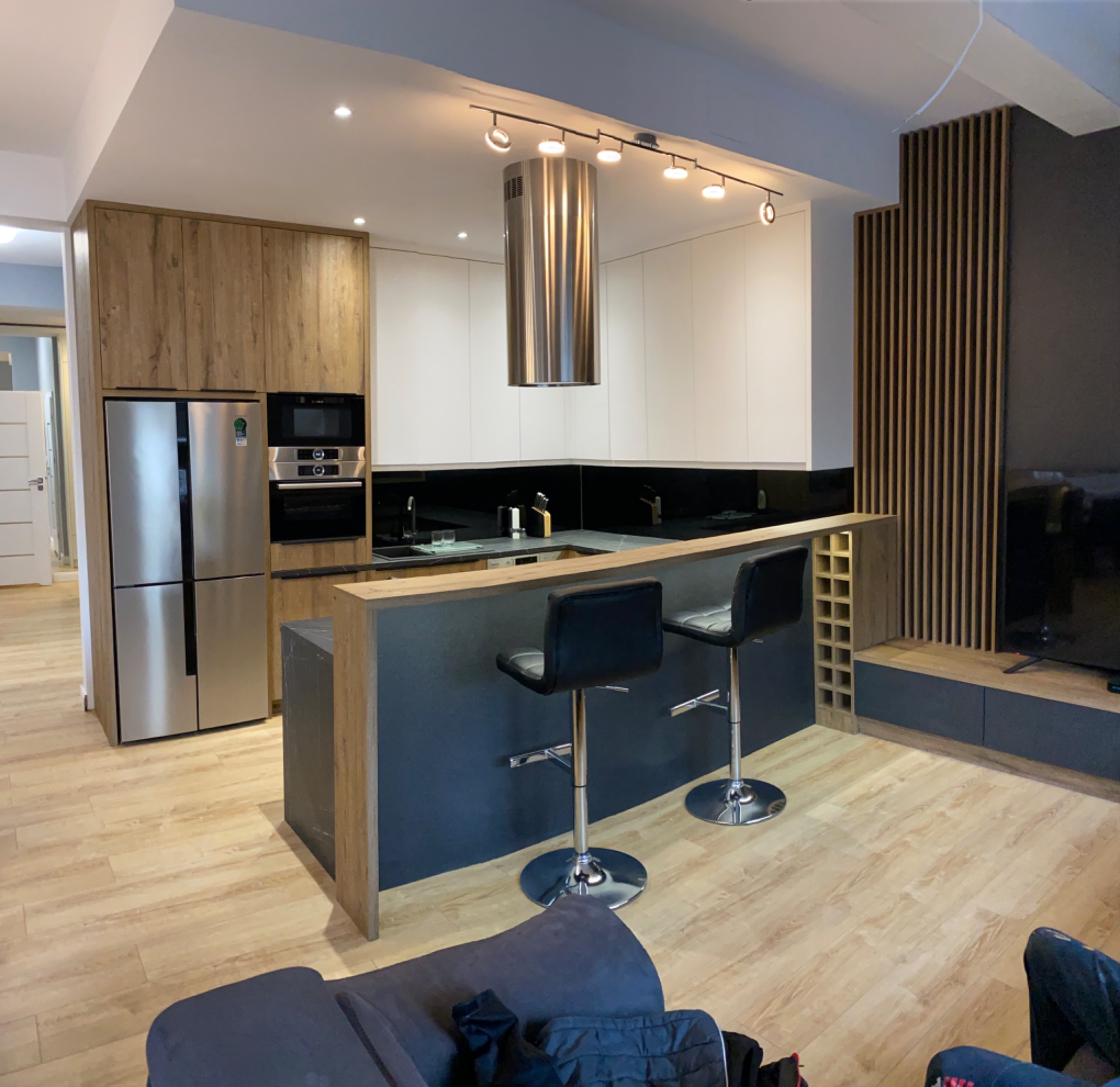 Nowoczesna kuchnia z wyspą, drewnianymi i białymi szafkami, stalowym okapem i czarnymi blatami, uzupełniona dwoma czarnymi hokerami