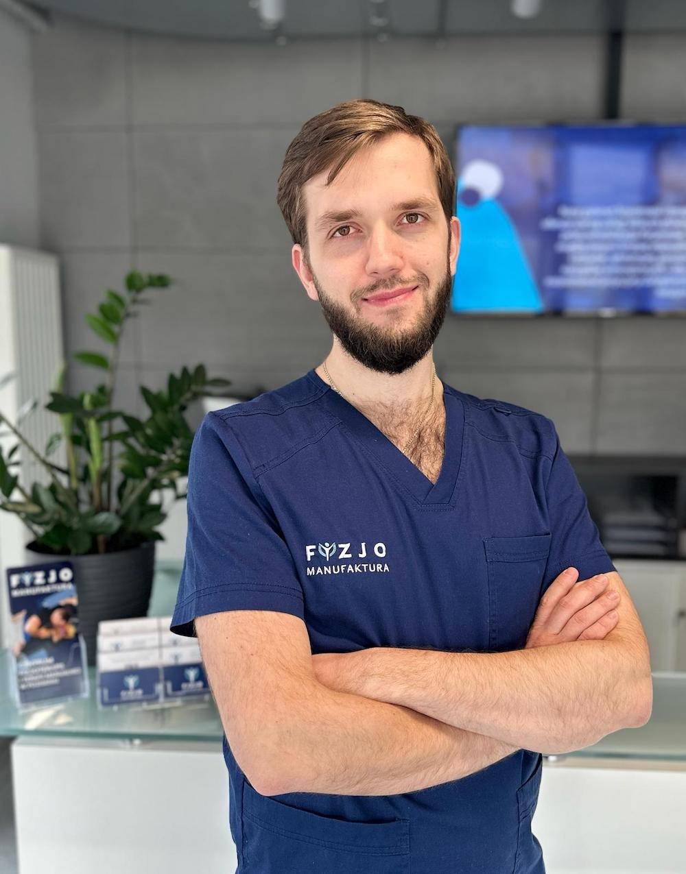 Uśmiechnięty mężczyzna w granatowym uniformie z logo 'Fyzjo Manufaktura', stojący na tle gabinetu z rośliną doniczkową i ulotkami na szklanym blacie.