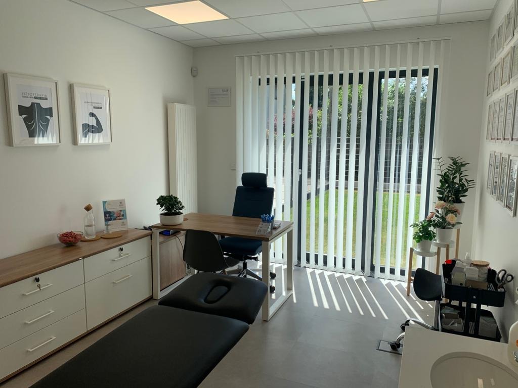 Jasne wnętrze gabinetu z kozetką do masażu, biurkiem i oknem z żaluzjami pionowymi, dekorowane roślinami i grafikami na ścianach.