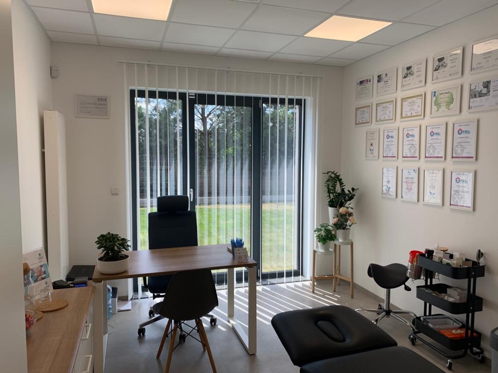 Jasne wnętrze gabinetu z leżanką do masażu, biurkiem, krzesłami i ścianą z certyfikatami, widok na ogród przez okno.