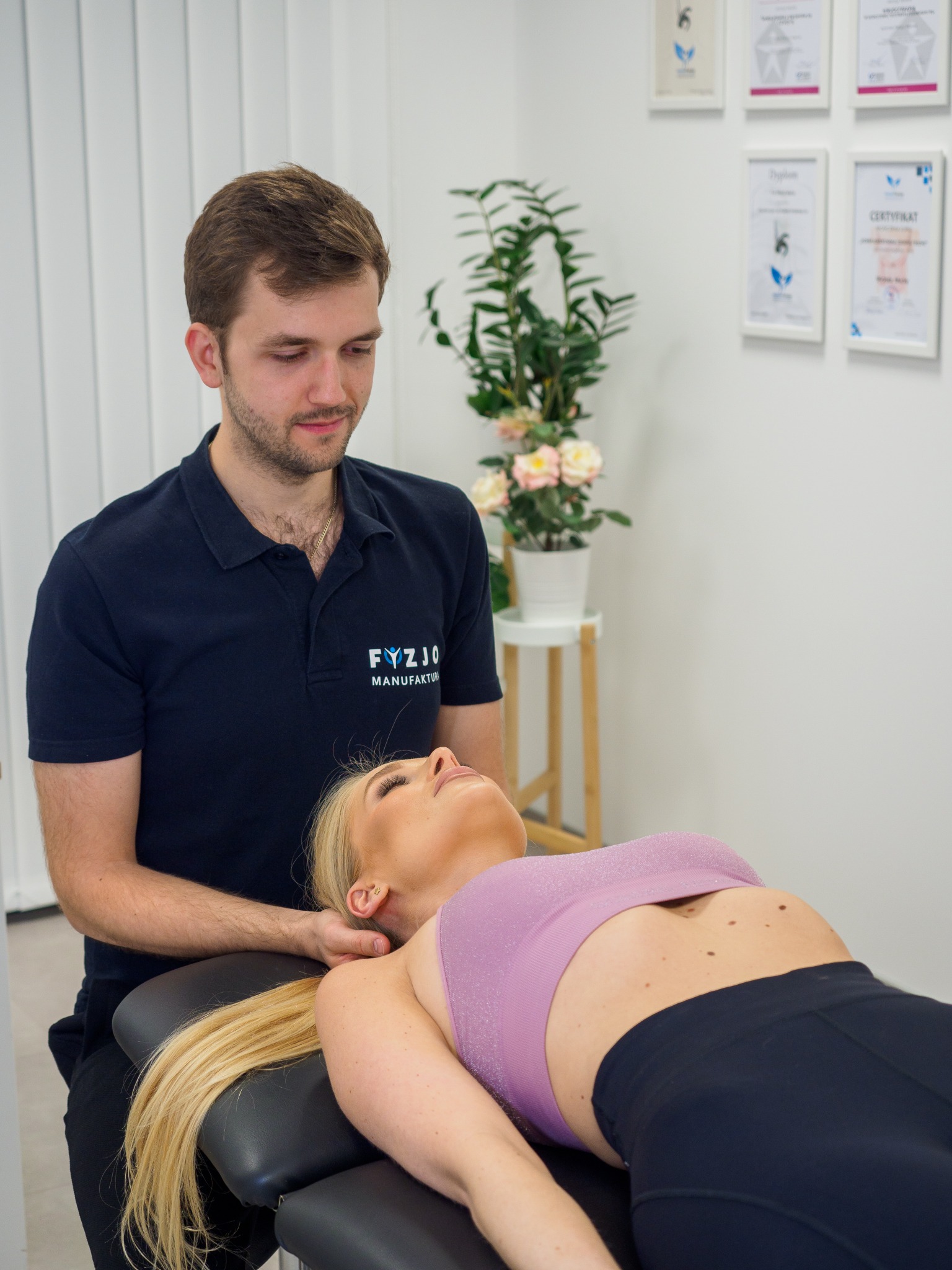 Fizjoterapeuta w granatowej koszulce z logo 'Fyzjo Manufaktura' delikatnie masuje szyję blondynki leżącej na czarnym stole do masażu w jasnym gabinecie, w tle widoczne dyplomy i kwiaty.